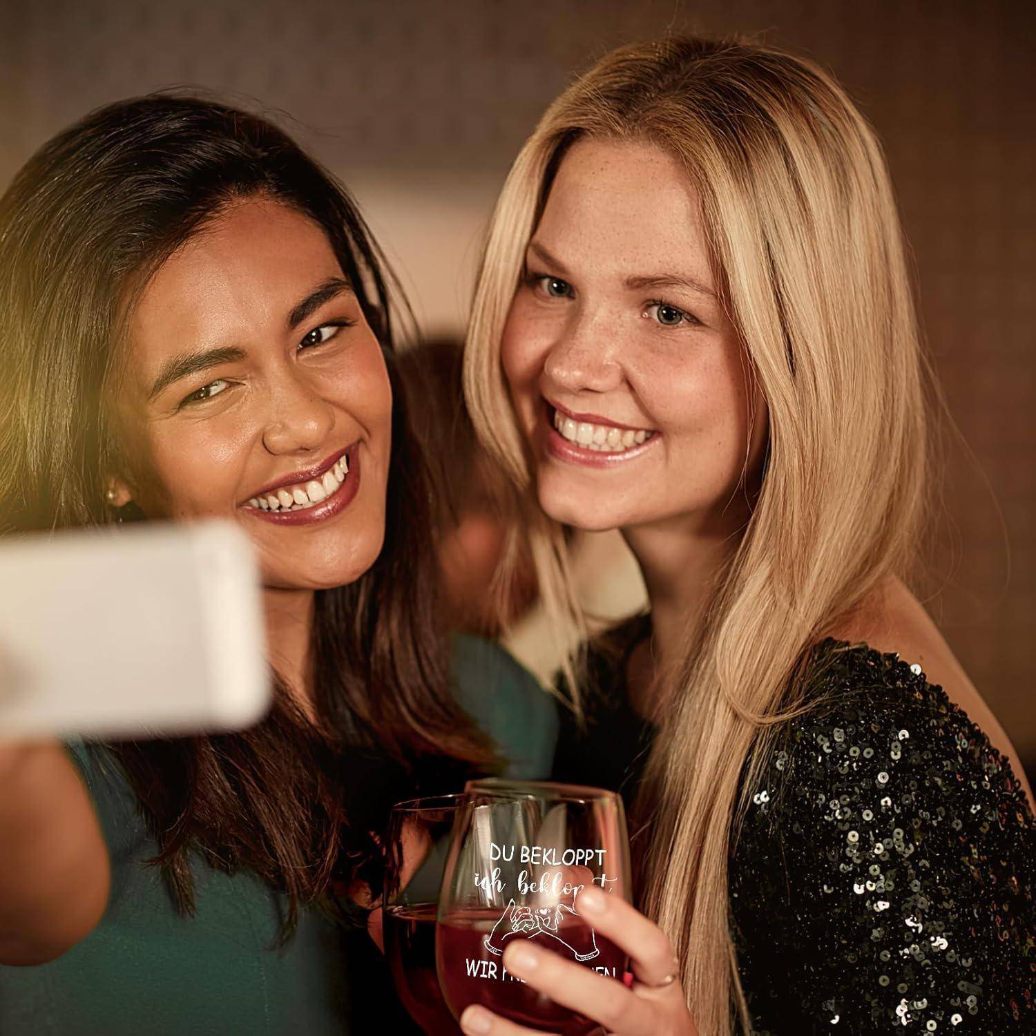 Zwei Frauen lächeln und machen ein Selfie. Eine hält ein Weinglas mit Text. Sie scheinen sich auf einer Indoor-Gesellschaftsveranstaltung zu befinden.