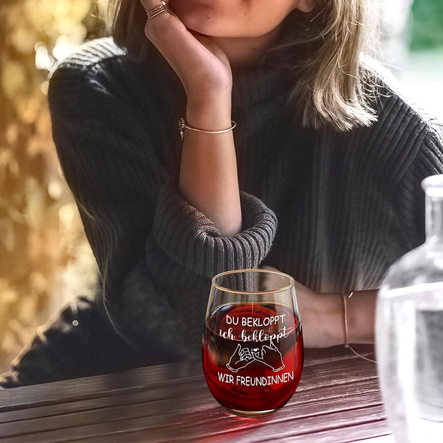 Eine Person in einem grauen Pullover, die ein Weinglas hält. Das Glas trägt den deutschen Text: 