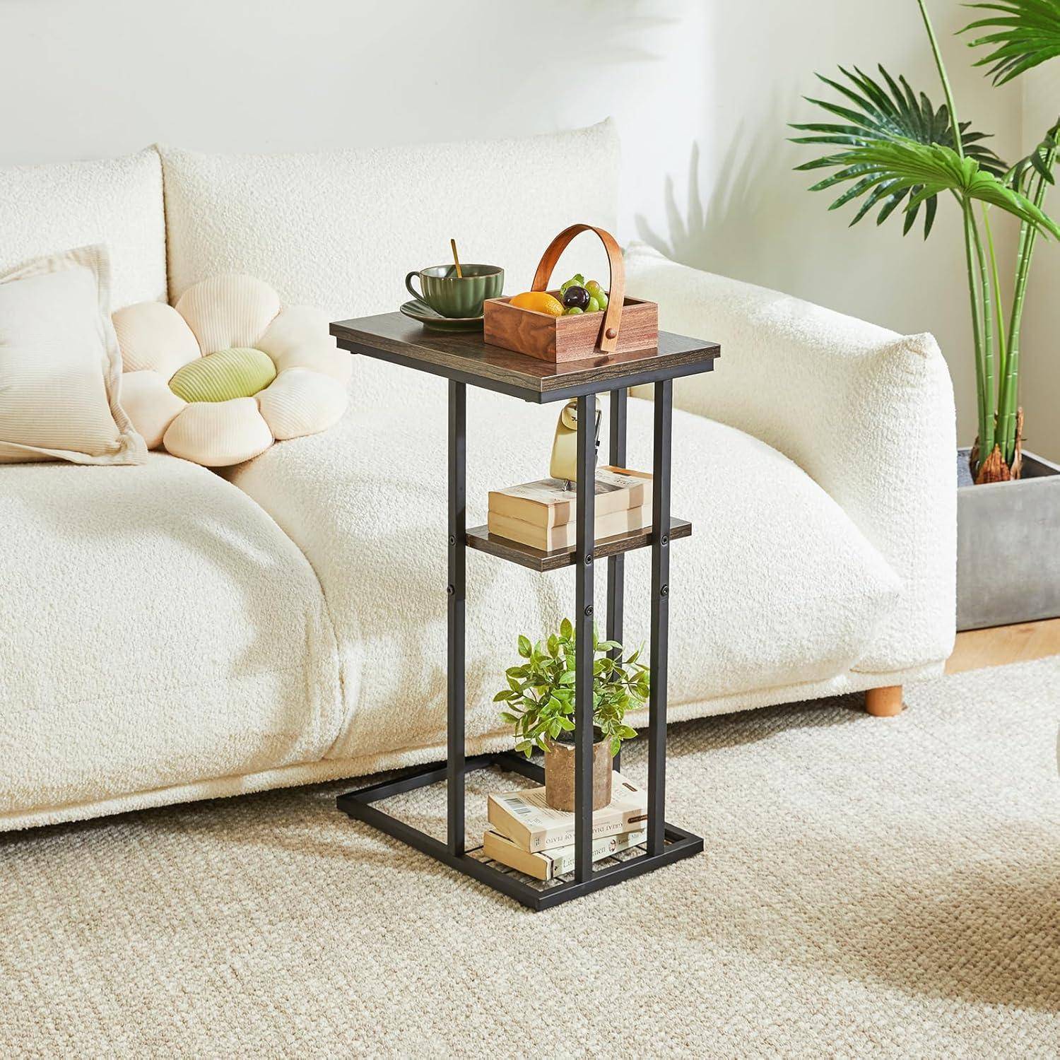 Ein kleiner Beistelltisch aus schwarzem Metall und Holz mit drei Ablagen, auf dem sich ein Obstkorb und eine Pflanze befinden, neben einem weißen Sofa in einem Wohnzimmer.