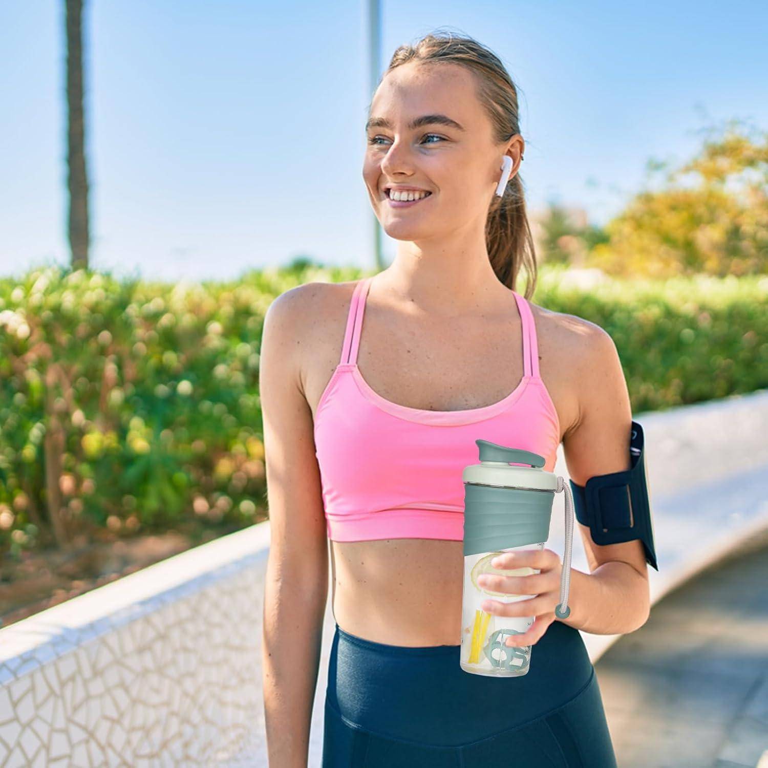Eine Joggerin in einem pinken Sport-BH und mit Ohrhörern hält eine Wasserflasche und lächelt an einem sonnigen Tag im Freien.