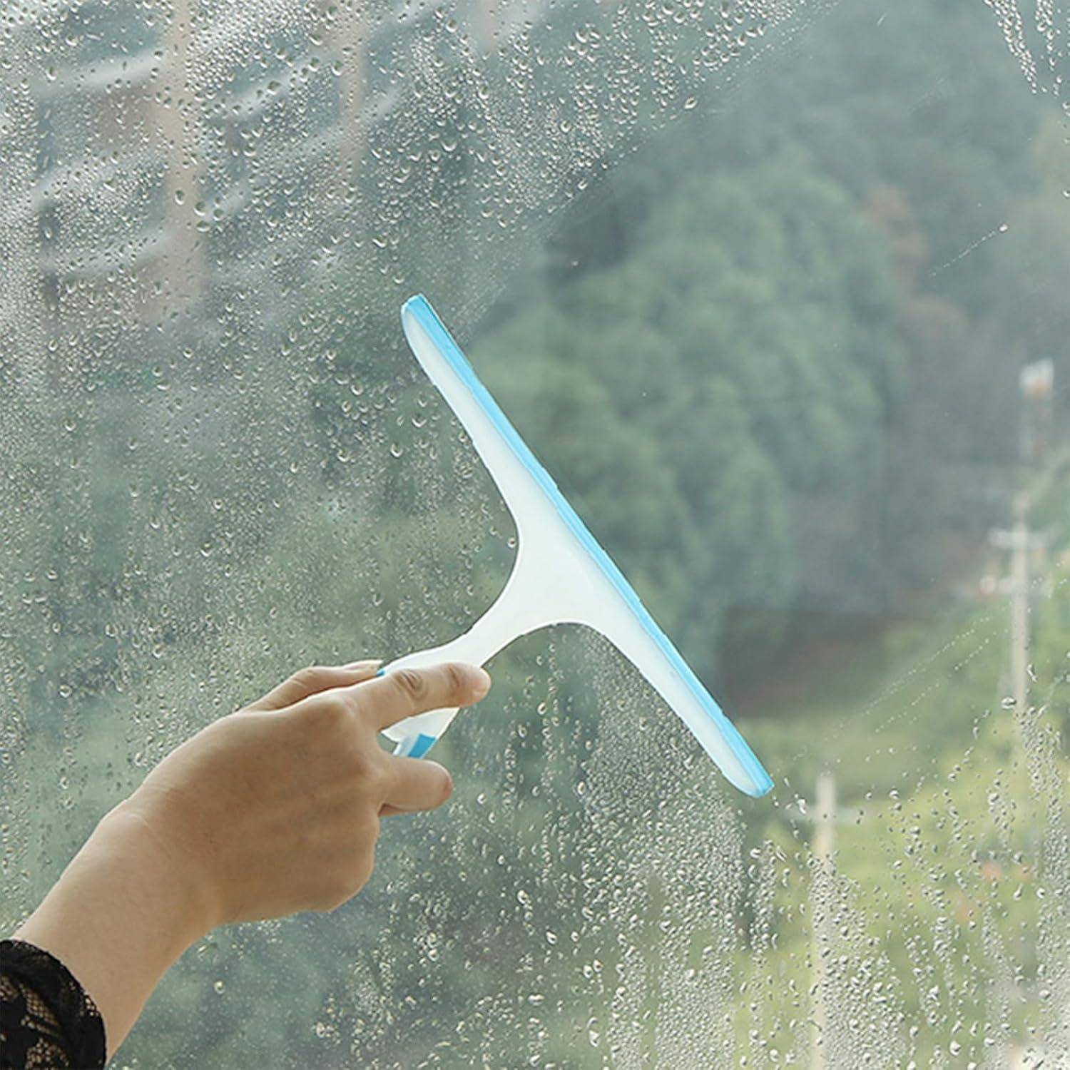 Eine Hand reinigt ein Fenster mit einem Abzieher und entfernt Wassertropfen. Verschwommene Außenkulisse mit Bäumen ist durch das Glas sichtbar.