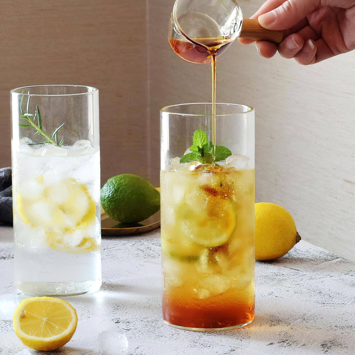 Zwei hohe Gläser mit Eistee, garniert mit Minze und Limette, neben einem Glas, das aus einer kleinen Kanne mit Tee gefüllt wird, auf einer Theke.