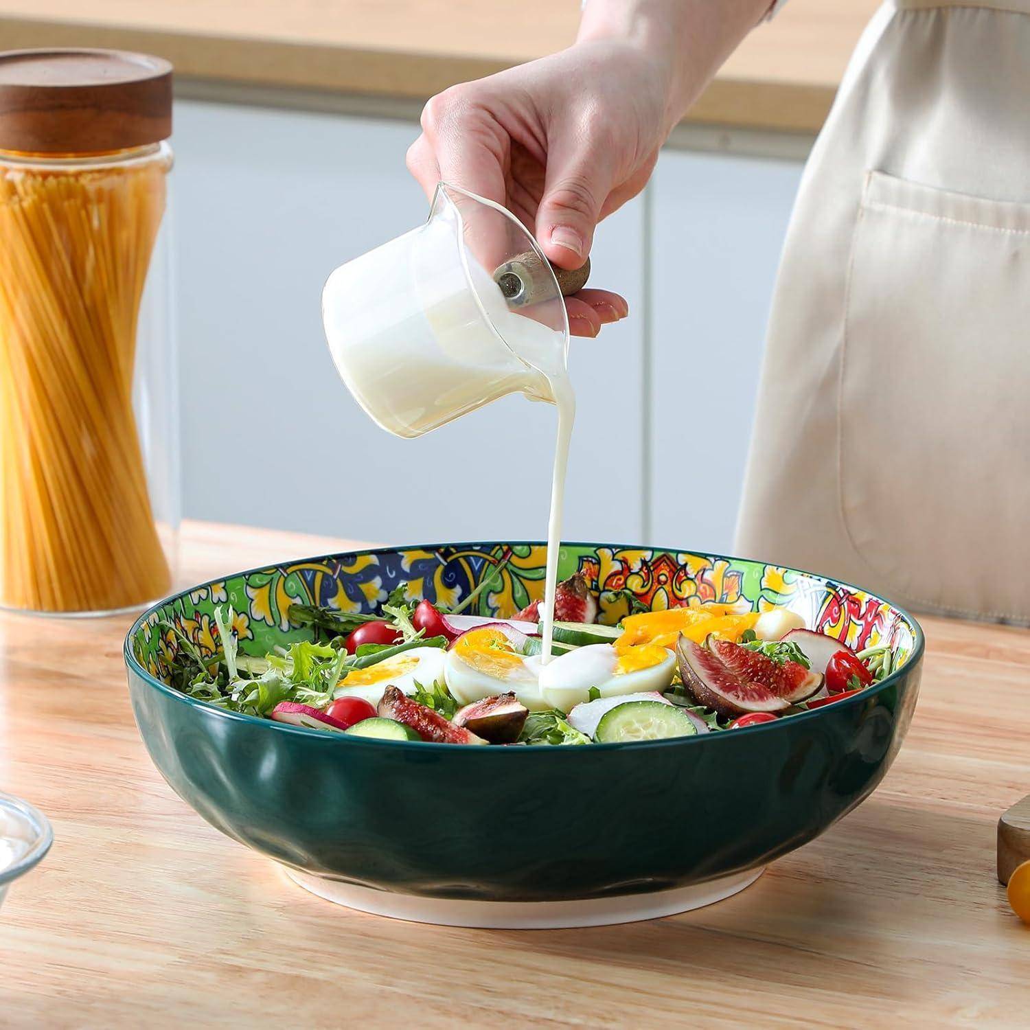 Eine Hand gießt Dressing über einen bunten Salat mit Eiern, Feigen und Tomaten in einer dekorativen Schüssel auf einer Küchenarbeitsplatte.