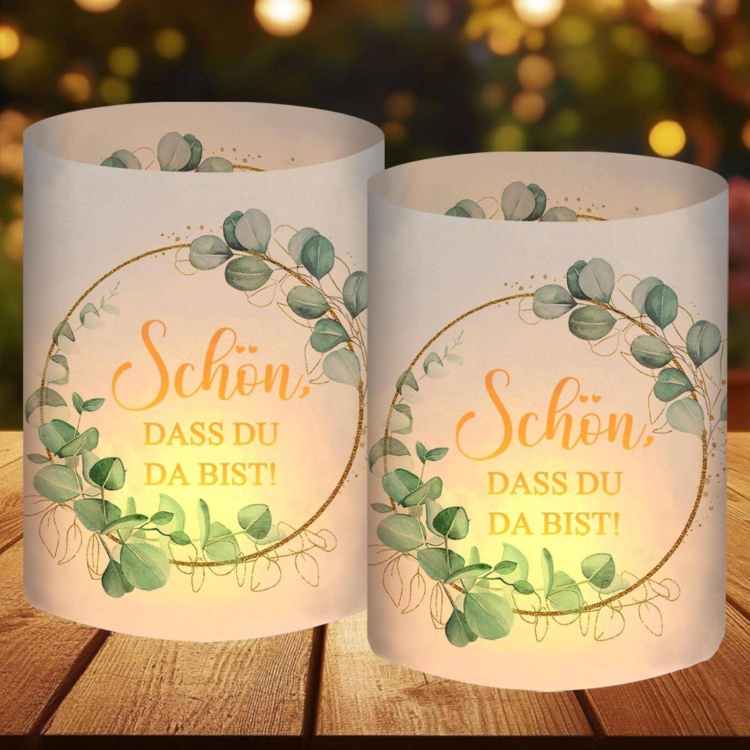 Zwei verzierte Kerzenhalter mit grünen Blättern und dem Text 'Schön, dass du da bist!' leuchten sanft vor einem unscharfen Hintergrund.
