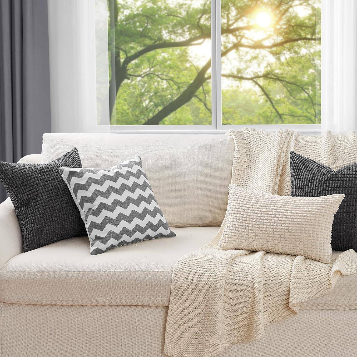 Ein gemütliches Wohnzimmer mit einem weißen Sofa, geschmückt mit Chevron- und strukturierten Kissen, einer weichen beigen Decke und einem Fensterblick auf einen sonnigen Tag.