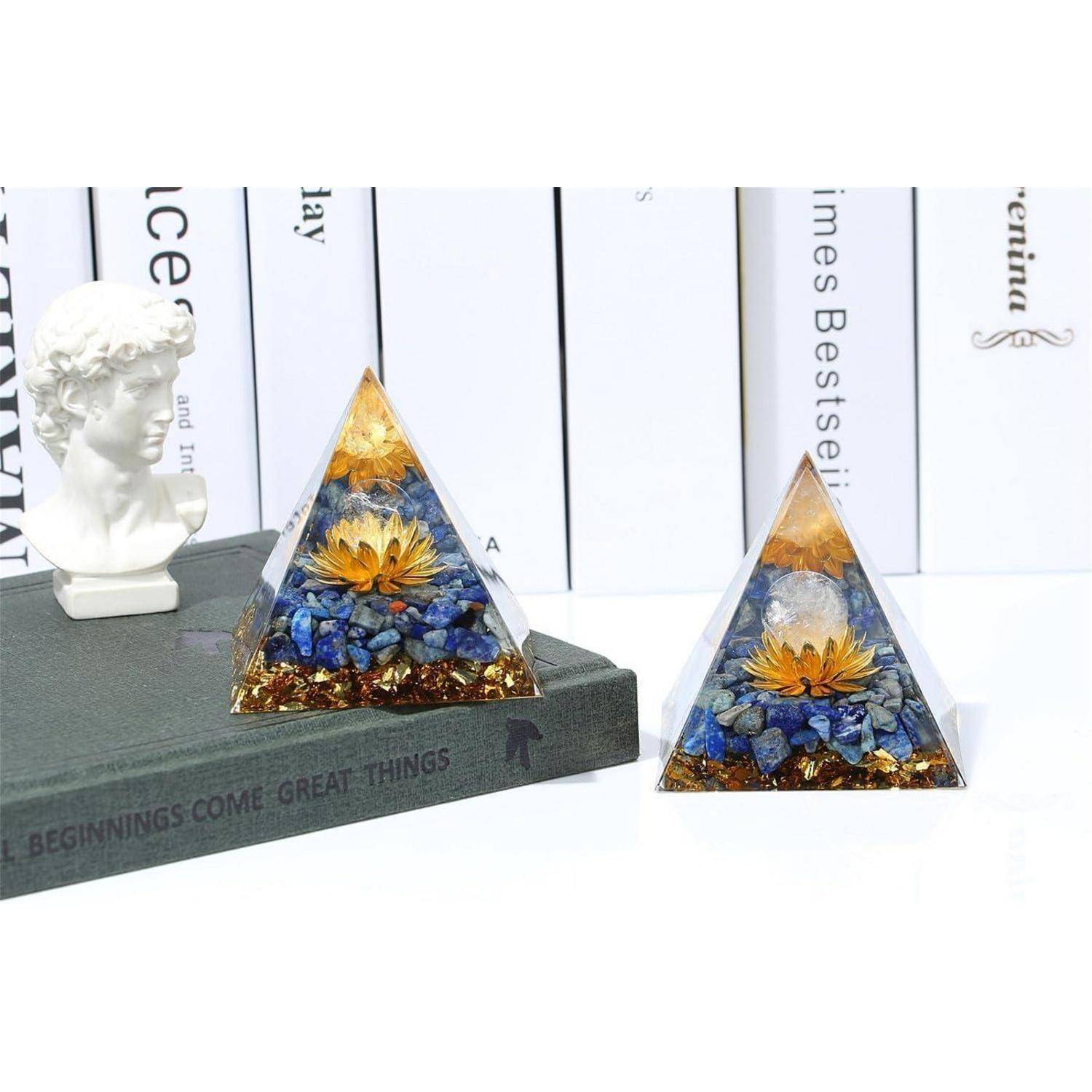 Zwei Kristallpyramiden stehen auf einem Buch neben einer kleinen Büste. Jede Pyramide enthält eine goldene Blume und blaue Steine im Inneren.