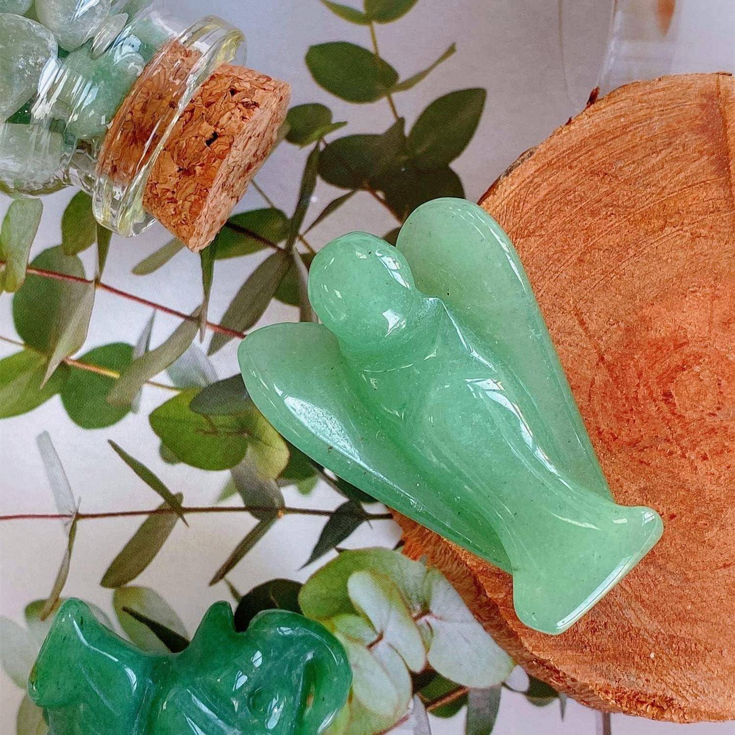 Eine geschnitzte grüne Engelsfigur aus Kristall ist auf einer Holzscheibe platziert, umgeben von grünen Blättern und einer kleinen, mit Korken verschlossenen Glasflasche.
