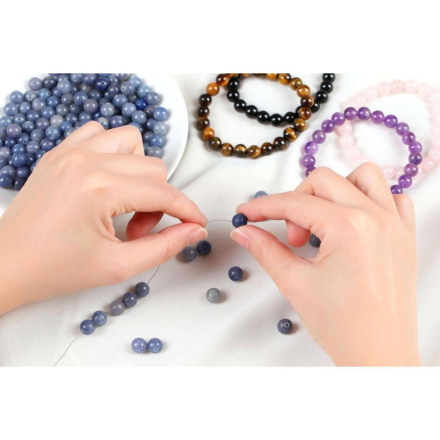 Hände, die blaue Perlen an einem Faden auffädeln; verschiedene Perlenarmbänder im Hintergrund, darunter braune, rosa und lila.