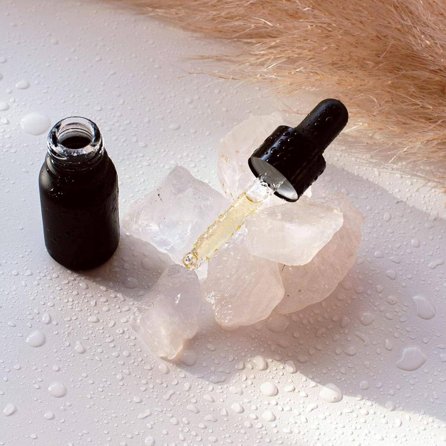 Eine schwarze Tropfflasche auf rosa Quarzkristallen mit ausgestreckter Flüssigkeitstropferpipette, umgeben von verstreuten Wassertropfen und einer weichen beigen Textur.