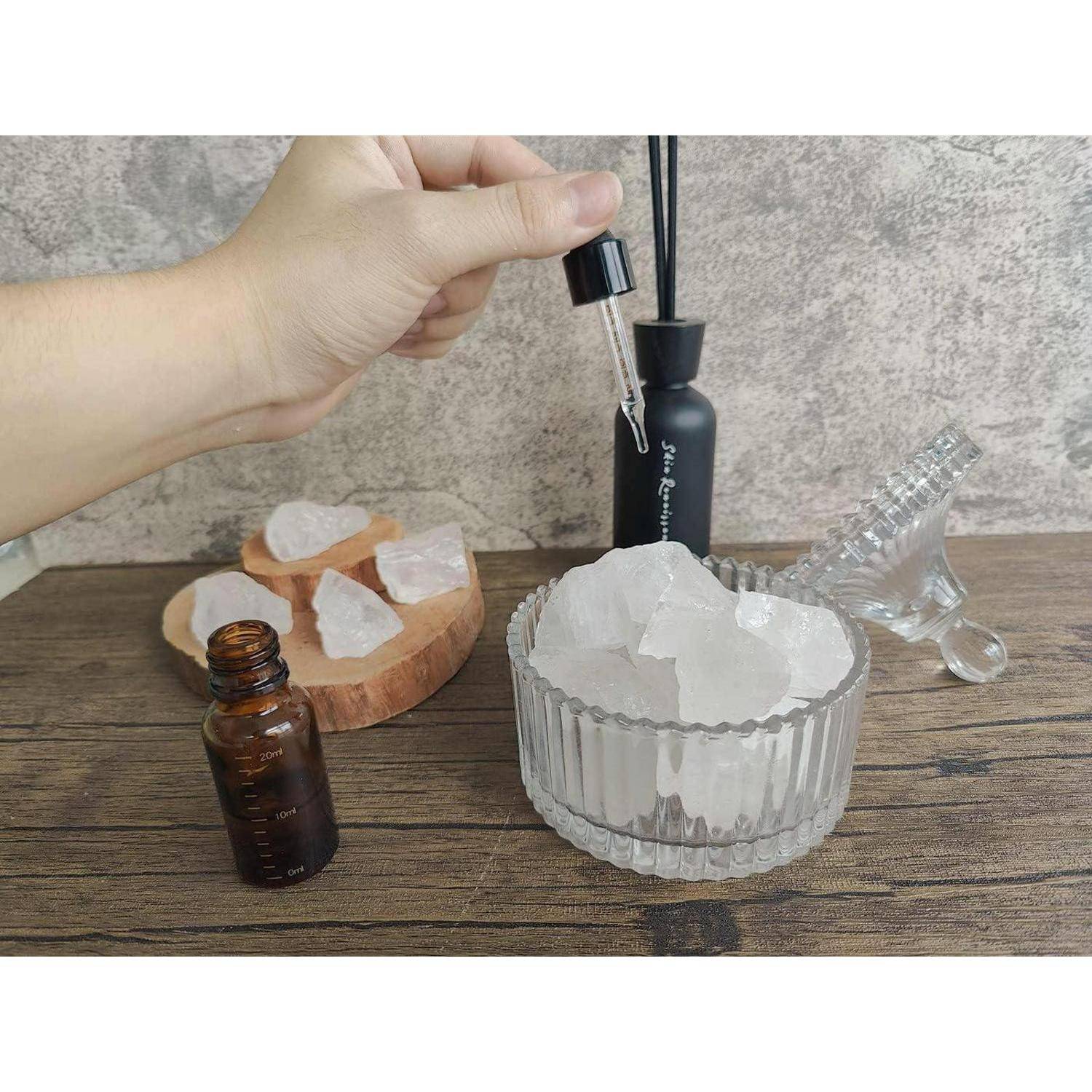 Eine Hand hält eine Pipette über ein Glas mit durchscheinenden Kristallen auf einer Holzoberfläche, mit einer kleinen braunen Flasche und dekorativen Gegenständen in der Nähe.