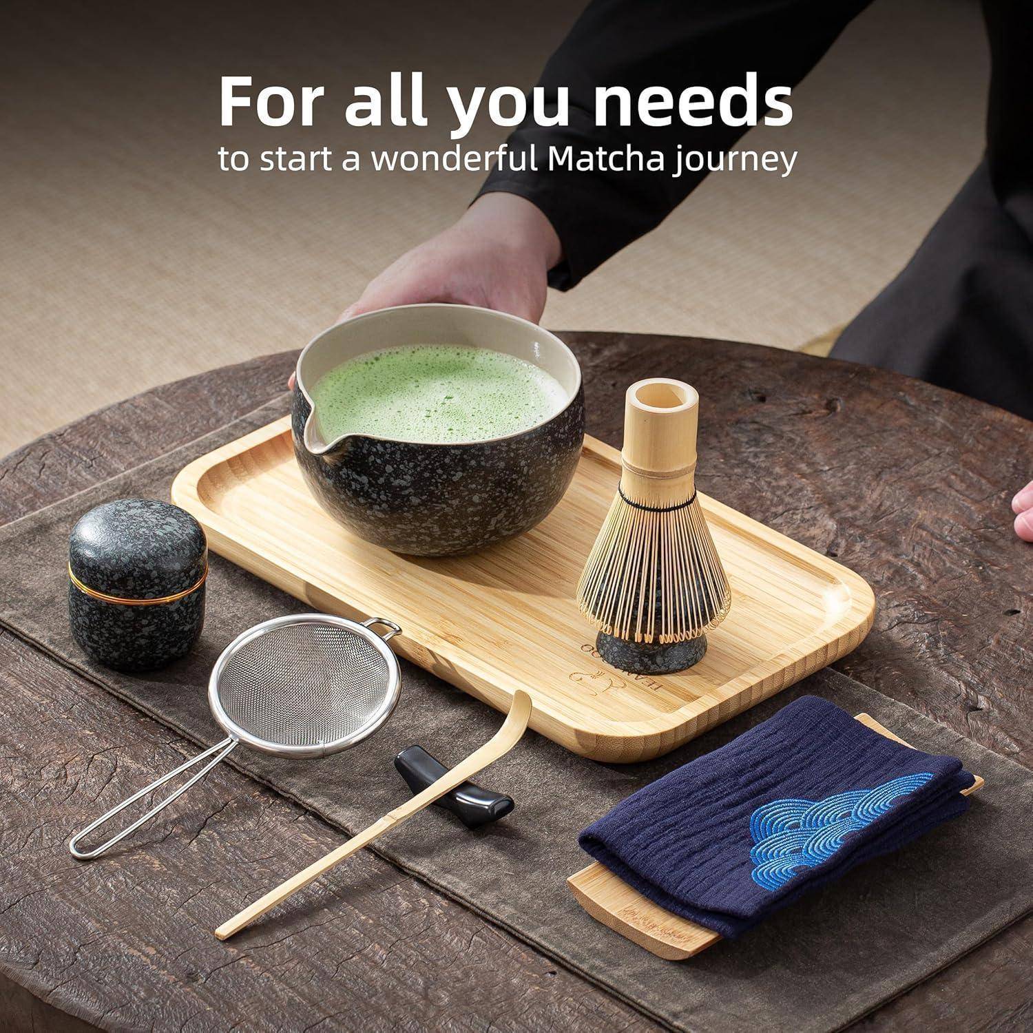 Matcha-Tee-Set auf einem Holztisch umfasst eine Schale mit schaumigem Matcha, einen Bambus-Schneebesen, ein Sieb, ein Gefäß und ein blaues Stoffserviette.