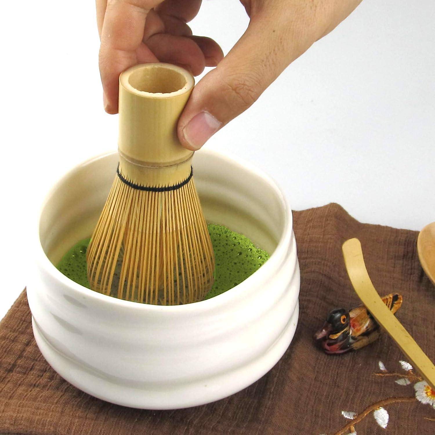 Eine Hand, die Matcha-Grüntee in einer weißen Schüssel mit einem Bambusbesen aufschlägt, platziert auf einer Holzoberfläche, mit einem dekorativen Tuch in der Nähe.
