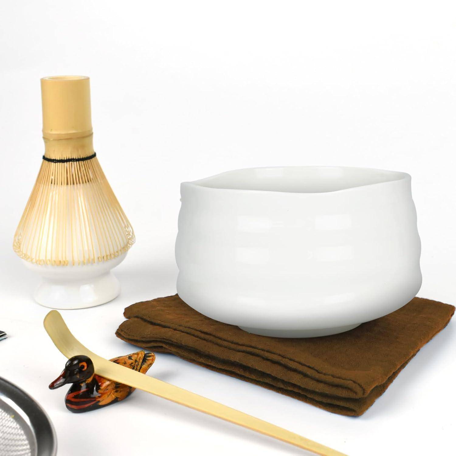 Ein japanisches Tee-Set mit einer weißen Schüssel, einem Bambusquirl, einem kleinen Holzstab und einer gefalteten braunen Serviette auf einer weißen Oberfläche.