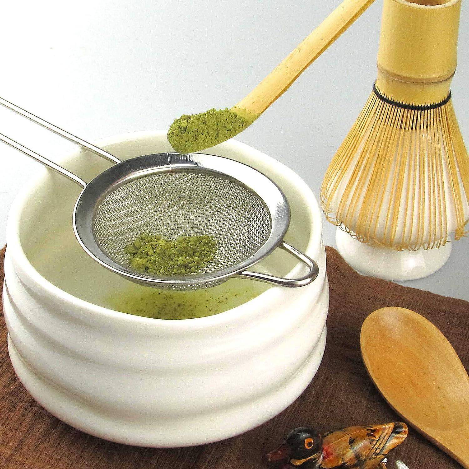 Eine traditionelle Teezeremonie-Anordnung zeigt eine weiße Schale mit einem Sieb, das grünes Matcha-Pulver enthält, einen Bambusbesen und einen Holzlöffel in der Nähe.