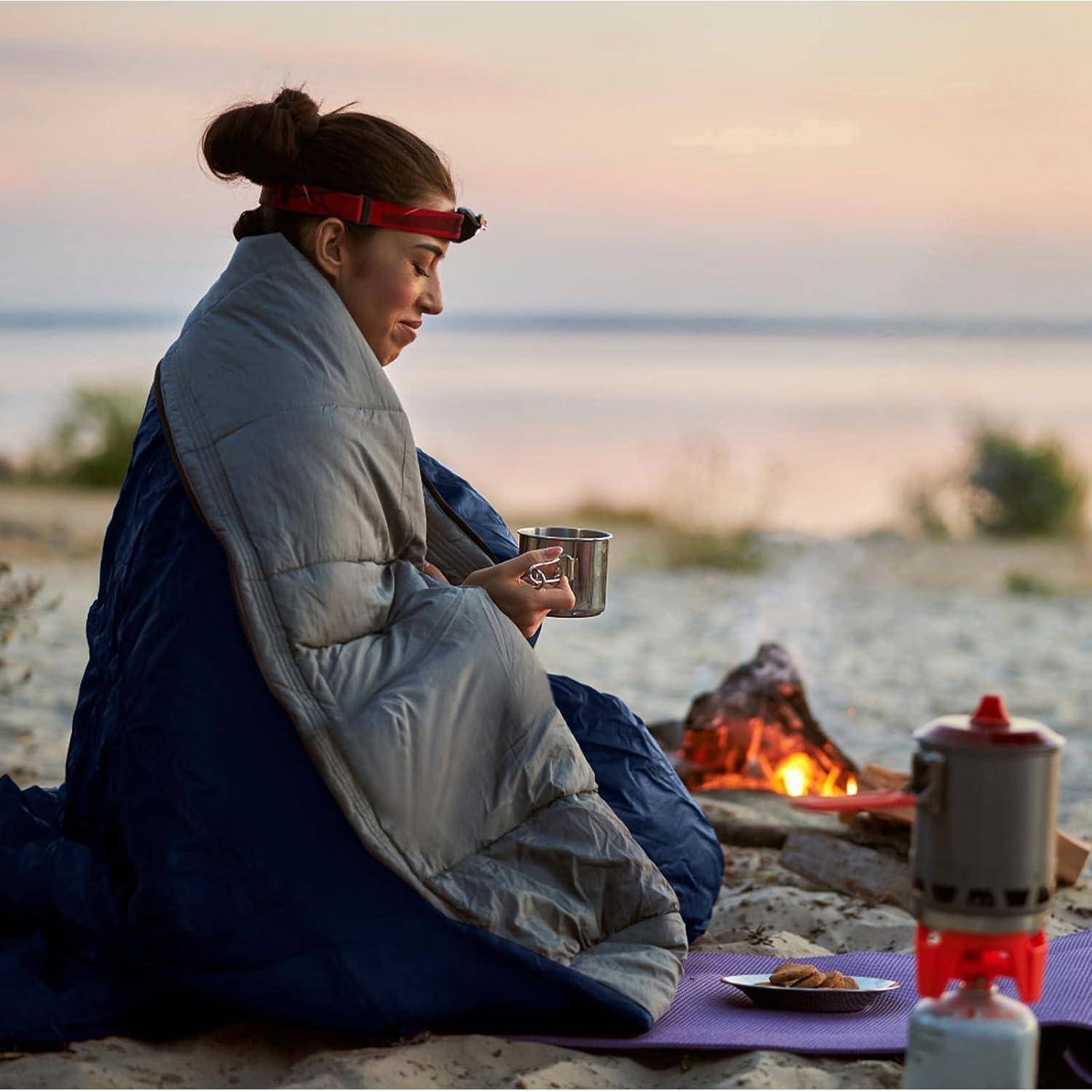 Eine Person sitzt in einen Schlafsack gehüllt am Strand bei Sonnenuntergang und hält eine Tasse. In der Nähe brennt ein kleines Lagerfeuer, und Campingausrüstung ist verstreut um sie herum.