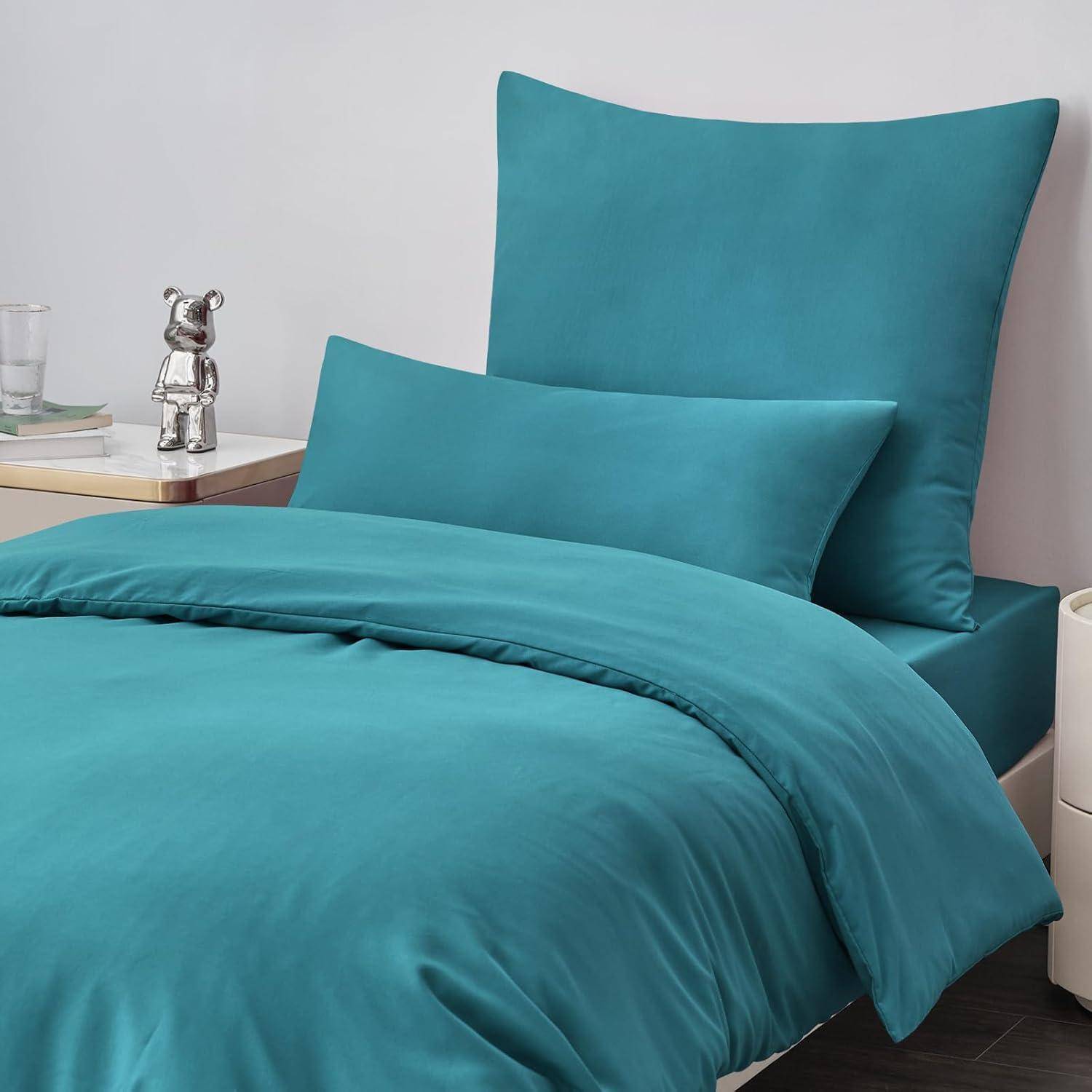 Ein ordentlich gemachtes Bett mit Petrolblau-Bettwäsche und Kissen in einer modernen Schlafzimmerumgebung, mit einer silbernen Teddybärfigur auf einem nahegelegenen Beistelltisch.