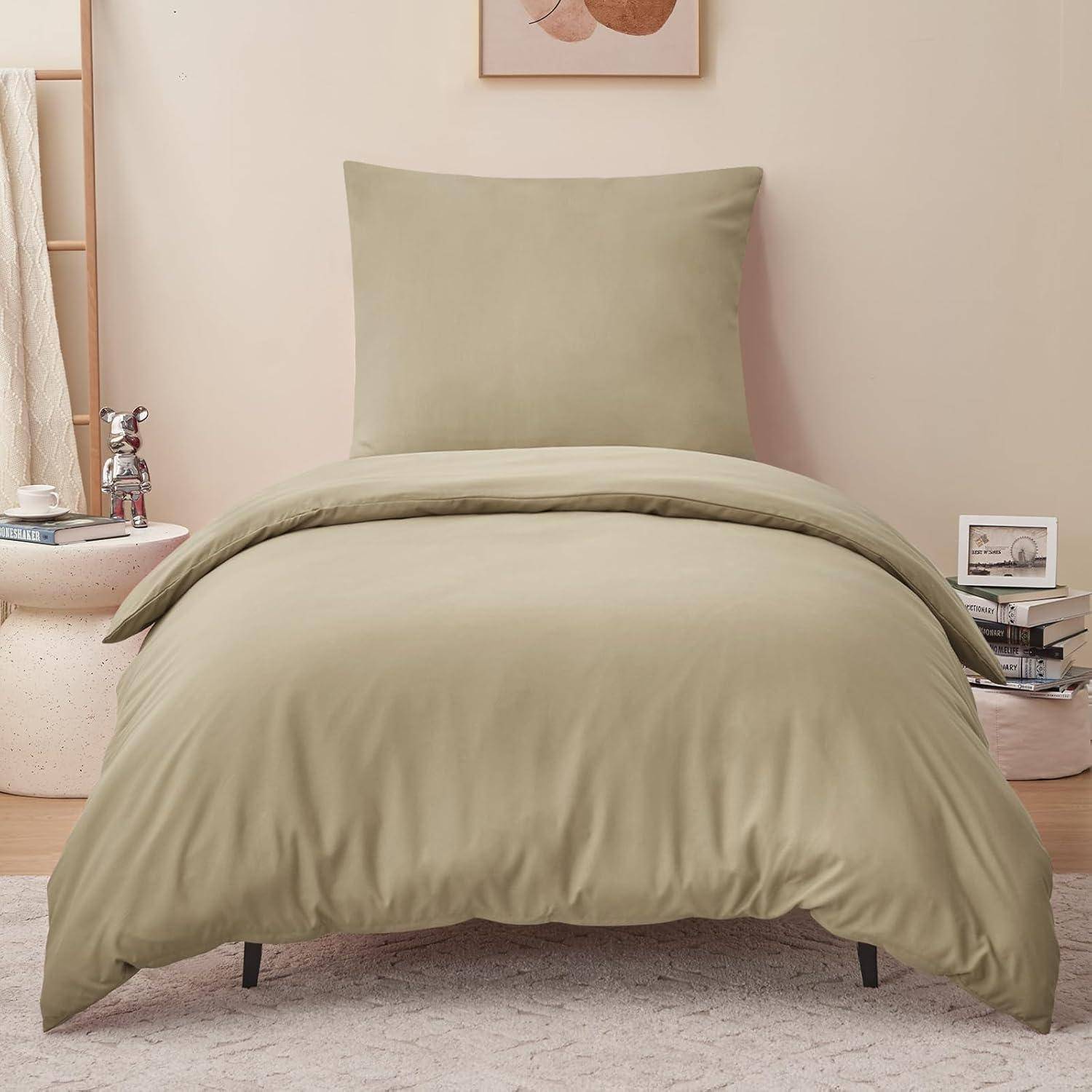 Ein ordentlich gemachtes Bett mit einem beigen Bettüberwurf vor einer hellgefärbten Wand. Ein kleiner Tisch mit Büchern und einer dekorativen Lampe befindet sich rechts davon.