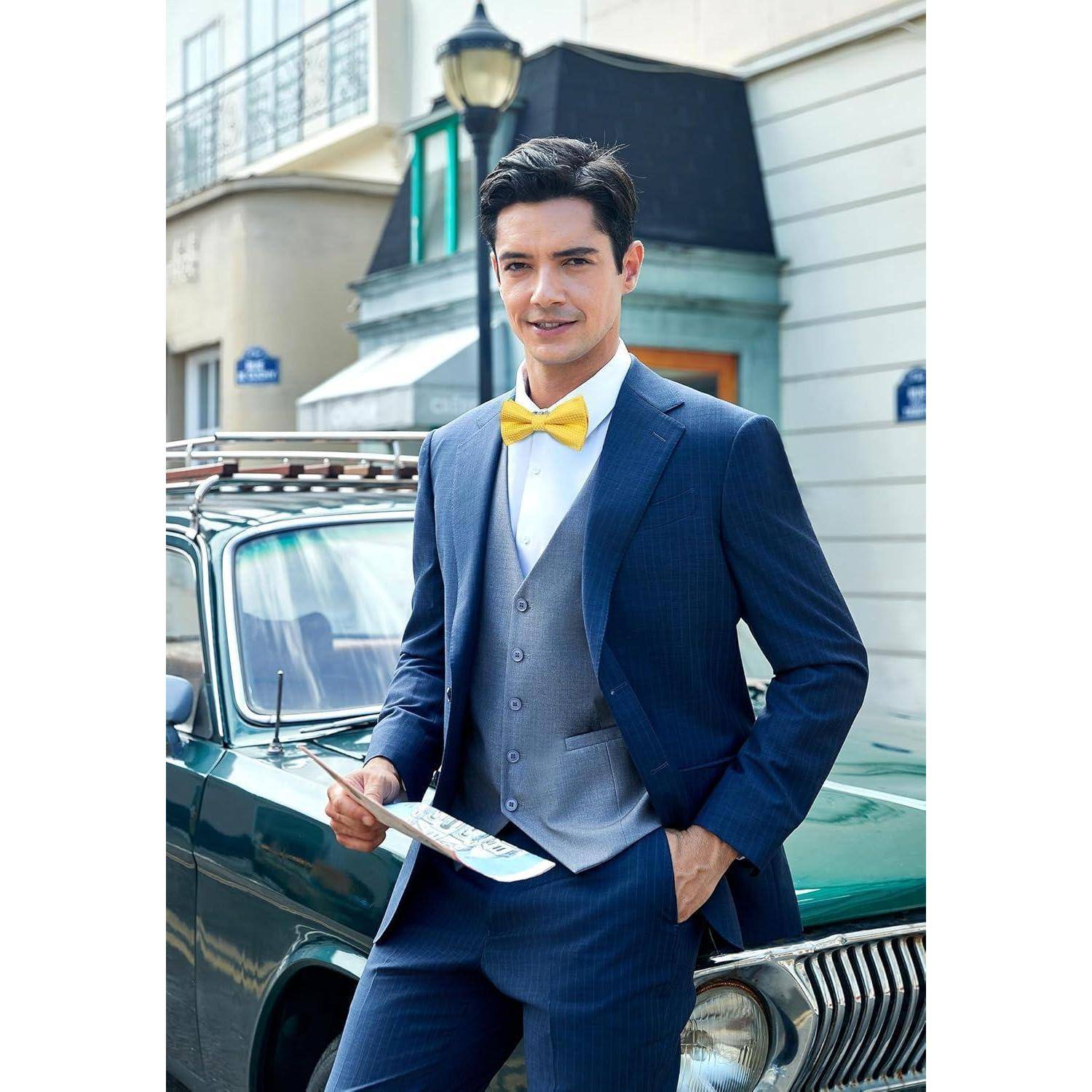 Ein Mann in einem blauen Anzug mit einer gelben Fliege steht vor einem Oldtimer und hält Papiere in der Hand, während er lächelnd auf einer Stadtstraße im Freien steht.