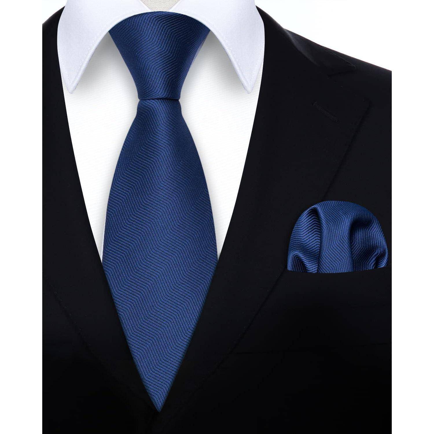 Eine Nahaufnahme einer Person in einem schwarzen Anzug, weißem Hemd und blauer Krawatte, mit einem gemusterten blauen Einstecktuch in der Jackentasche.