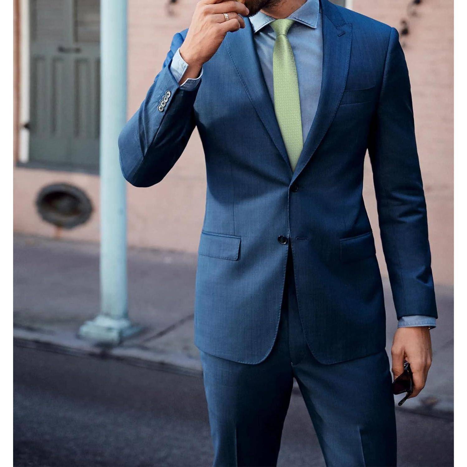 Eine Person in einem blauen Anzug mit einer hellgrünen Krawatte steht auf einem Stadtbürgersteig und hält ein Telefon. Der Hintergrund zeigt ein Gebäude mit einer grünen Tür.