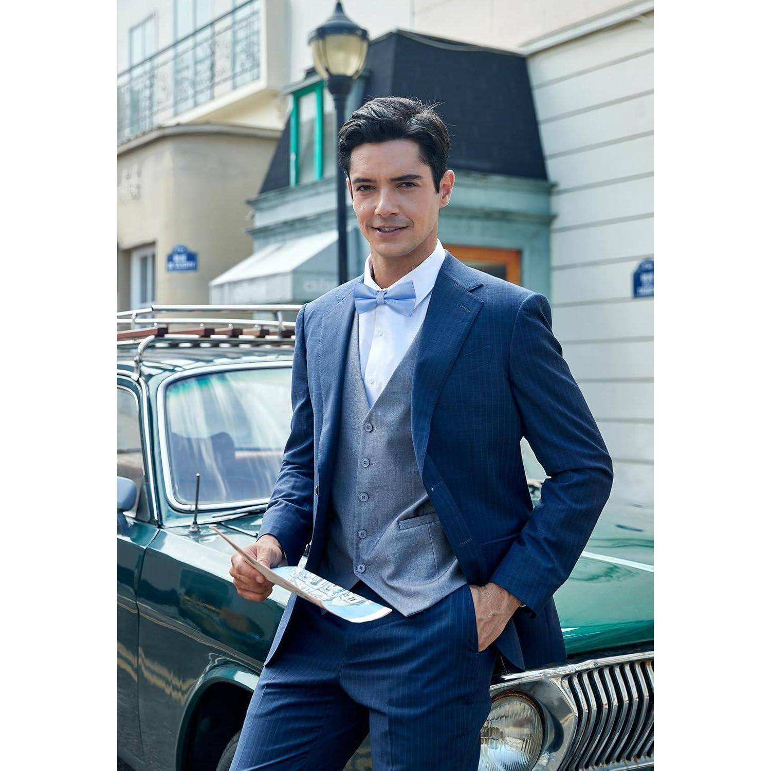 Ein Mann in einem blauen Anzug lächelt, während er neben einem Oldtimer auf einer Straße steht und eine Karte hält. Urbane Umgebung mit Gebäuden im Hintergrund.