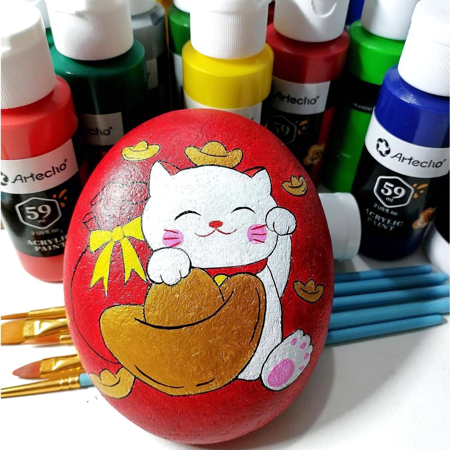 Ein bemalter Stein zeigt eine weiße Katze mit einer gelben Schleife und goldenen Details, umgeben von Acrylfarbenflaschen und Pinseln.