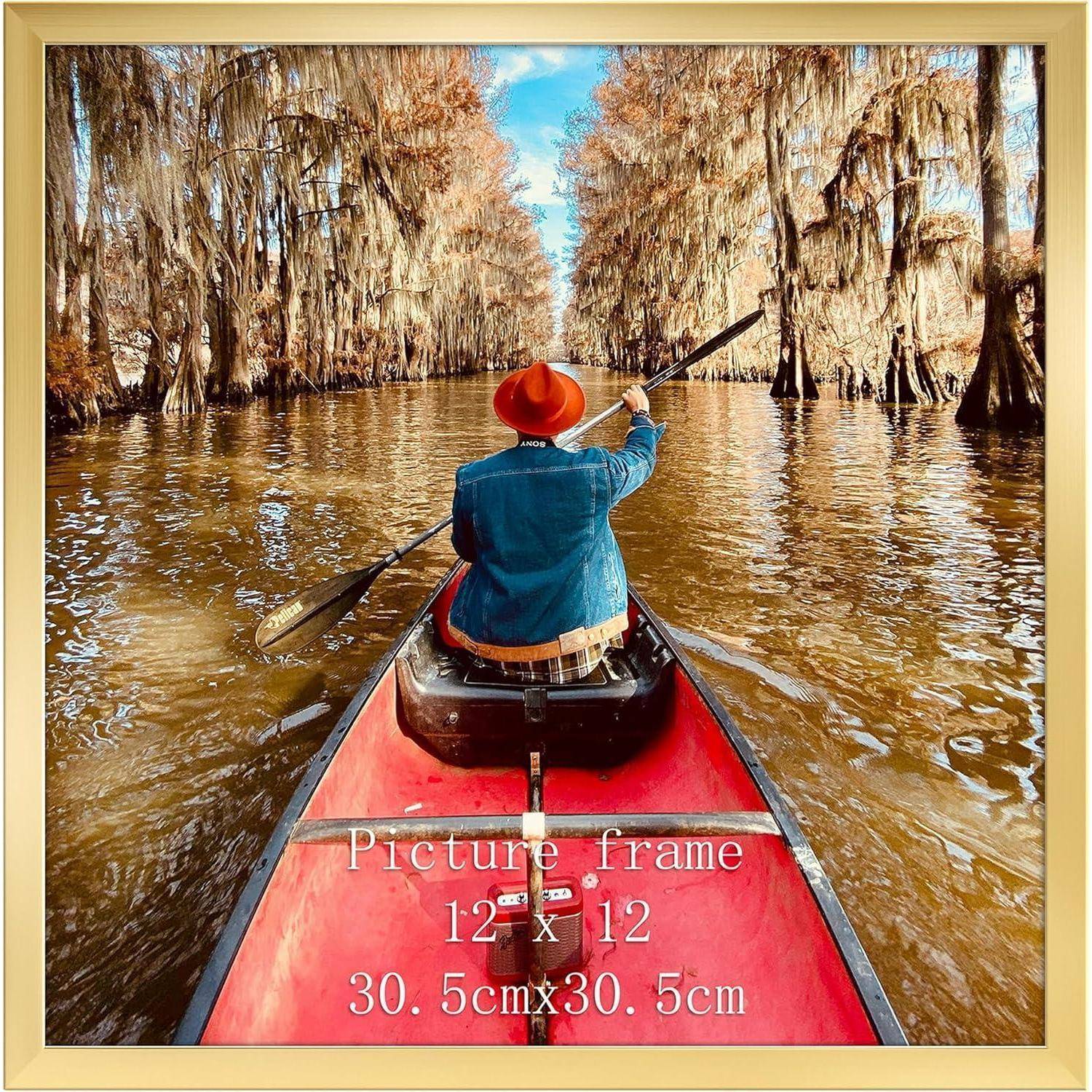 Eine Person mit roter Mütze paddelt ein Kanu durch einen von Zypressen gesäumten Wasserweg unter blauem Himmel. Das Bild ist in einem Bilderrahmen mit den Maßen '30,5 cm x 30,5 cm'.