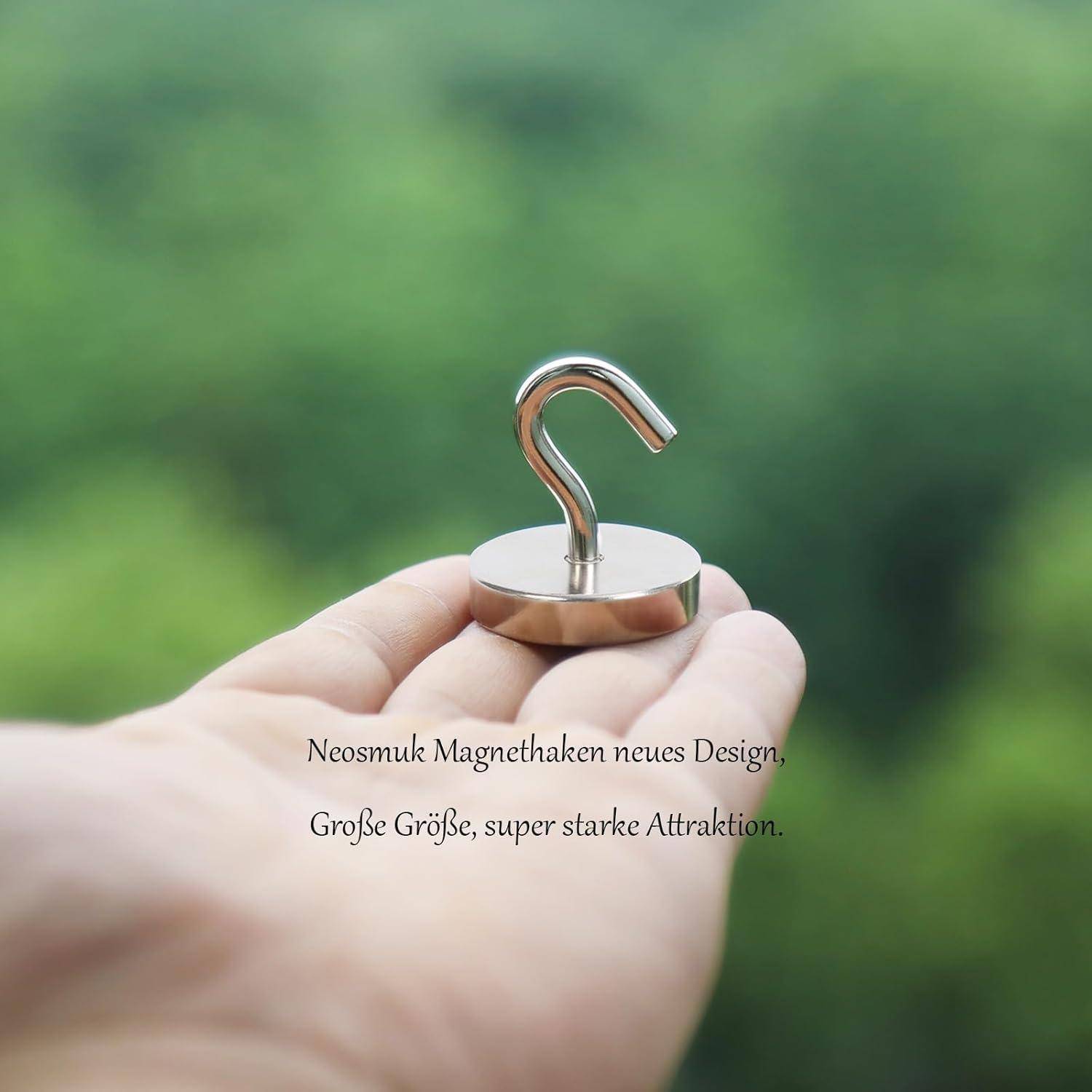 Ein Handgriff hält einen Metallhaken vor einem unscharfen grünen Hintergrund. Der Text auf Deutsch lautet: 'Neosmuk Magnethaken neues Design. Große Größe, super-starke Attraktion.