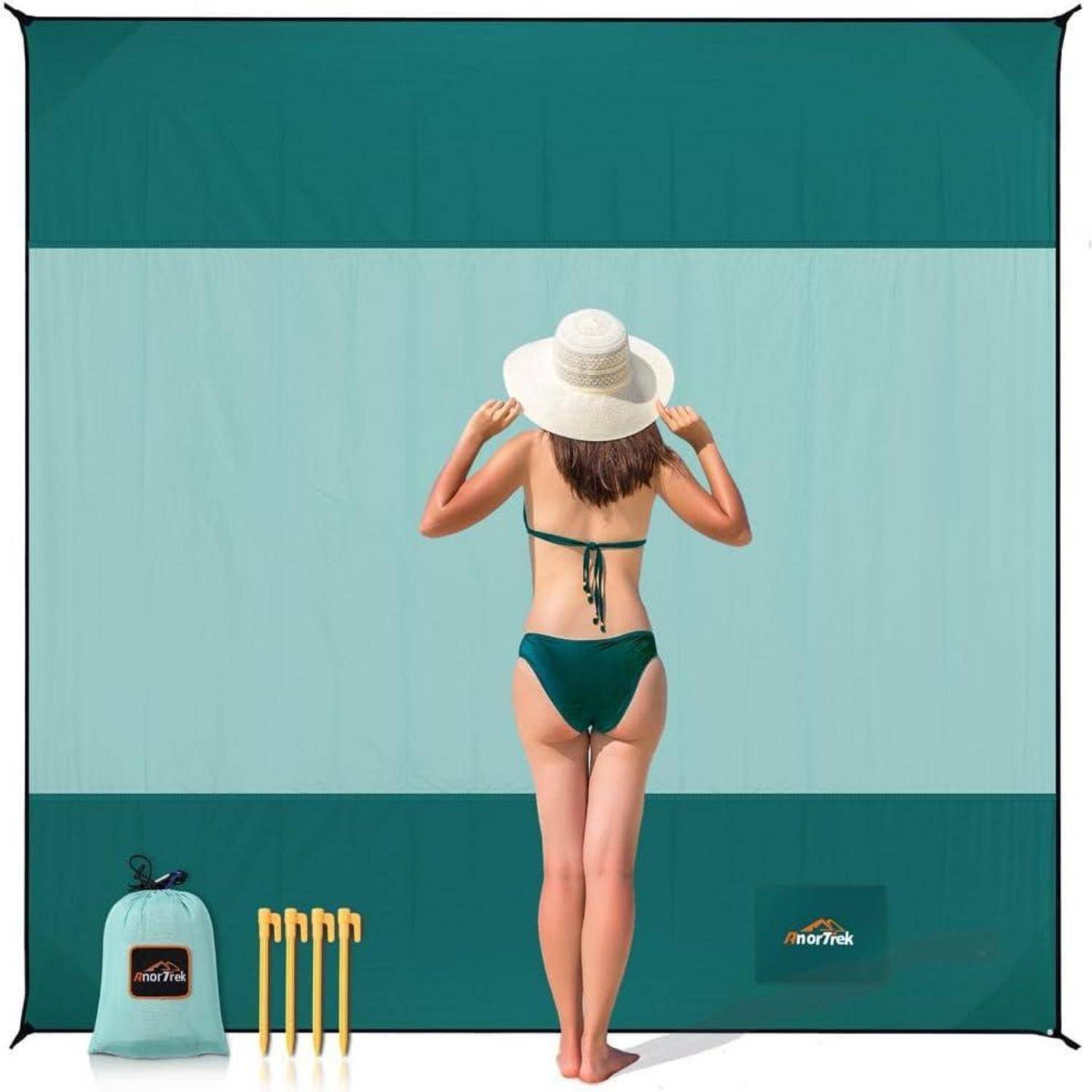 Eine Frau in einem grünen Bikini hält einen Sonnenhut und steht vor einer großen türkisfarbenen Stranddecke. Vier Pflöcke und eine Tragetasche befinden sich im Sand.