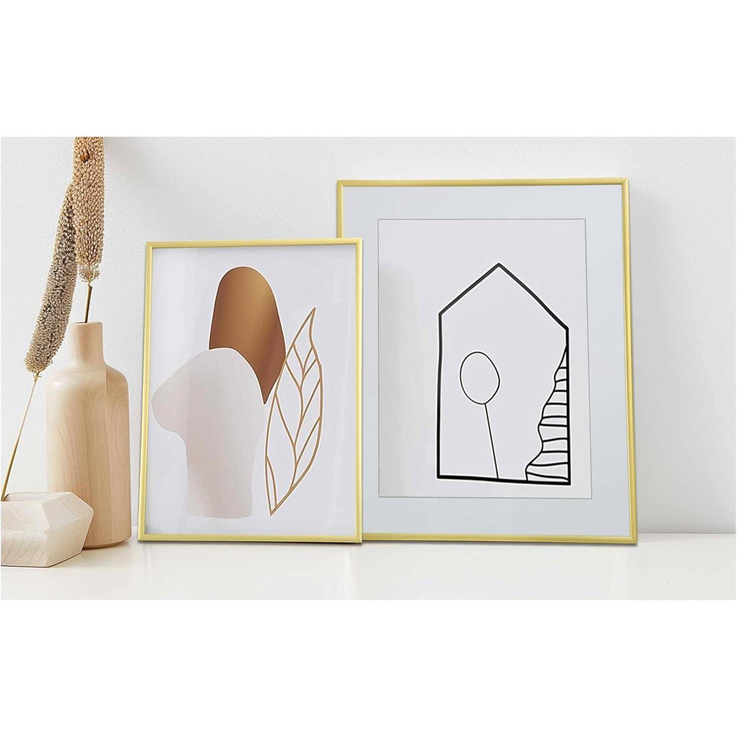 Zwei gerahmte abstrakte Kunstwerke auf einem Regal: eines mit einem minimalistischen Blatt- und Formdesign, das andere mit einer Hausumriss- und Baumdarstellung.