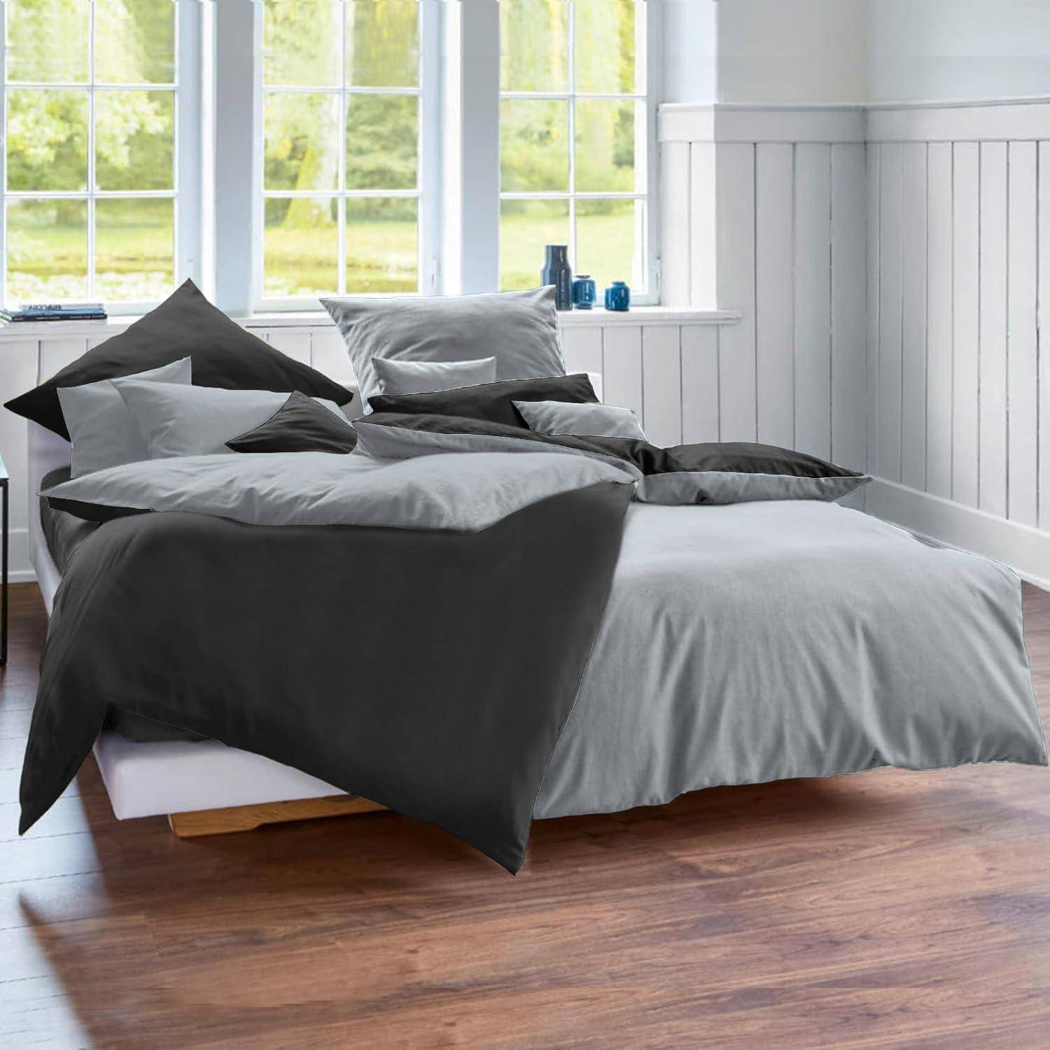 Ein ordentlich gemachtes Bett mit grau und anthrazitfarbener Bettwäsche steht in einem hellen Raum mit großen Fenstern und Holzboden.