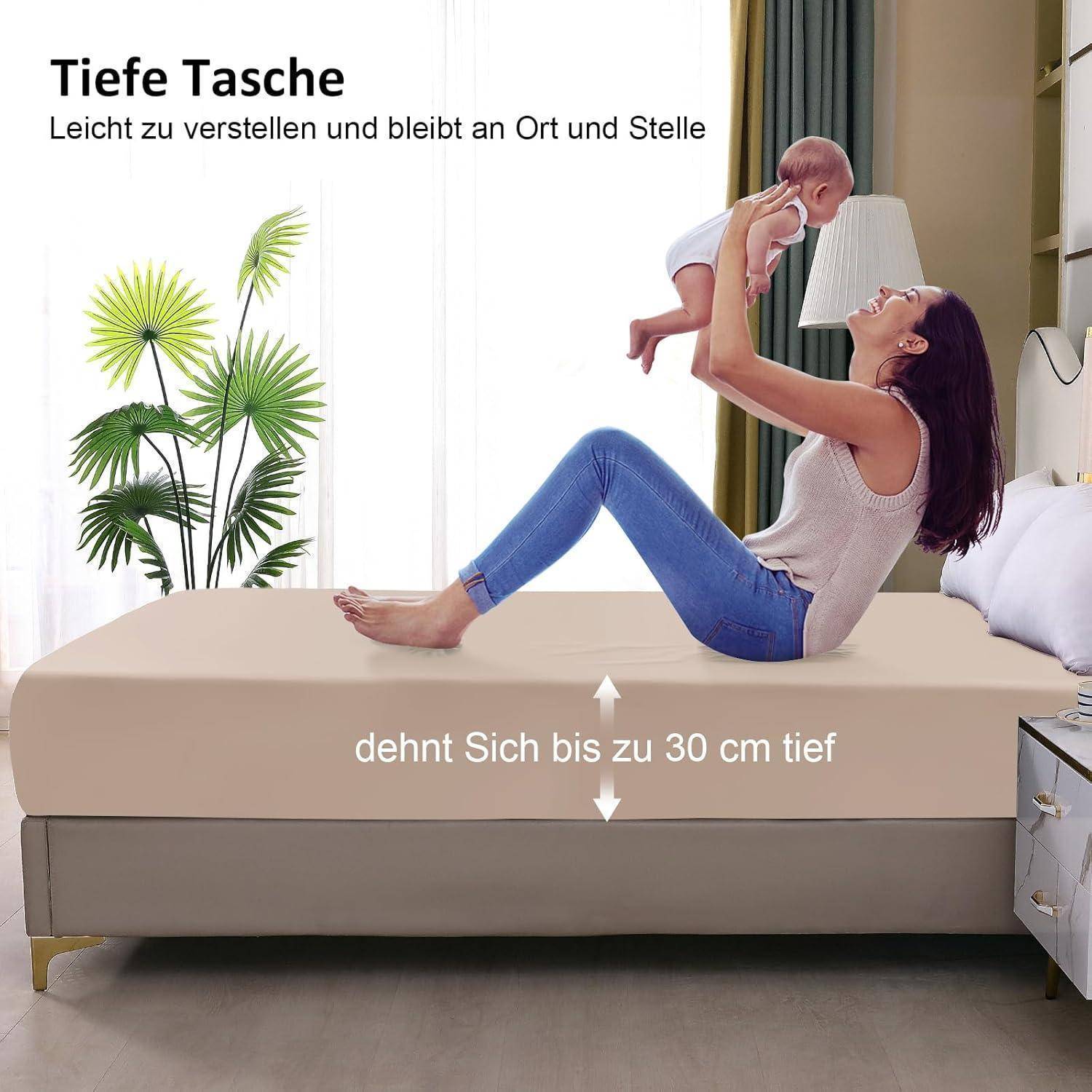 Eine Frau liegt auf einem beigen Bett und hält ein Baby in die Höhe, wobei der Bettlaken mit einer tiefen Tasche von bis zu 30 cm gezeigt wird. Text auf Deutsch beschreibt die Passform.