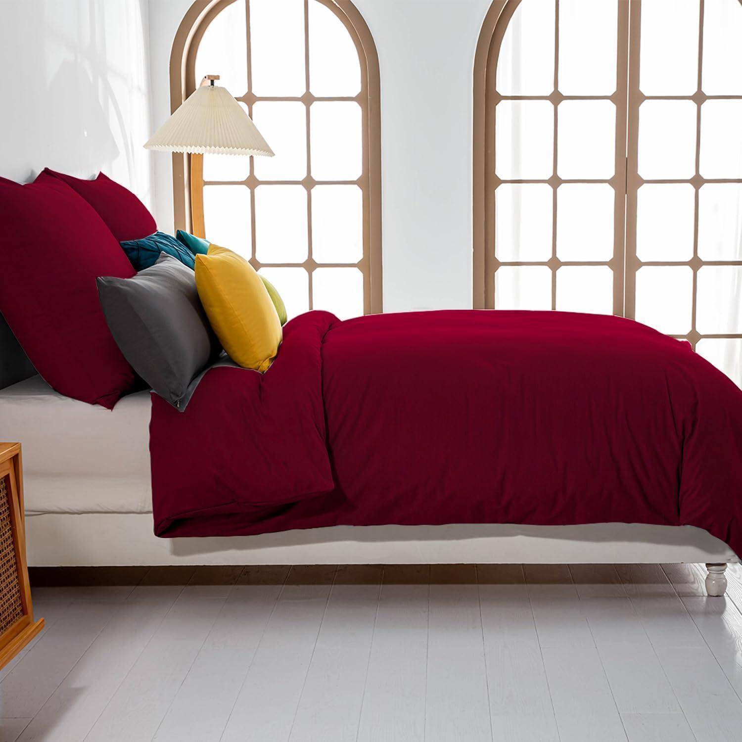 Ein gemütliches Schlafzimmer mit einem eleganten Bett mit Holzrahmen, bordeauxroter Tagesdecke, verschiedenen Kissen und einer Lampe, vor hohen Bogenfenstern.