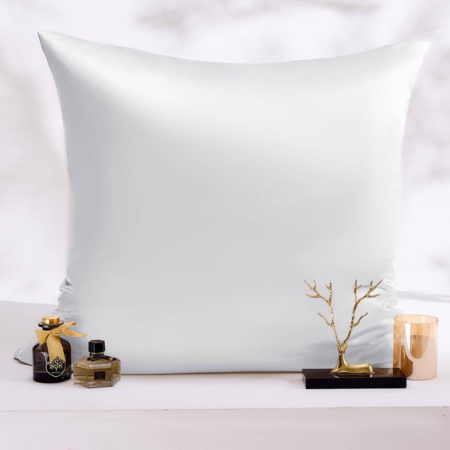 Ein großes weißes Kissen, zentriert zwischen zwei Parfümflaschen und einer kleinen goldenen Baumschmuck auf einer weißen Oberfläche, die eine minimalistische Ästhetik erzeugt.