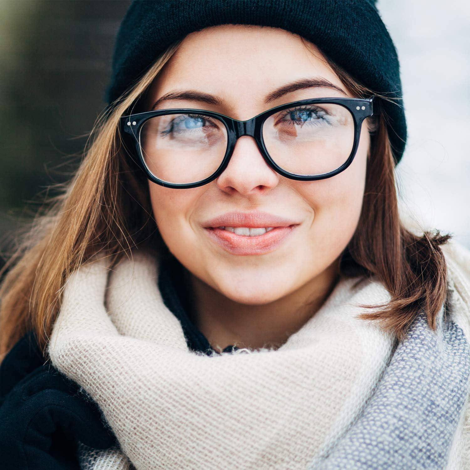 Eine Person mit langen Haaren, die eine Brille und eine schwarze Beanie trägt, lächelt sanft. Sie ist in einen gemütlichen Schal gehüllt, was auf kaltes Wetter hindeutet.