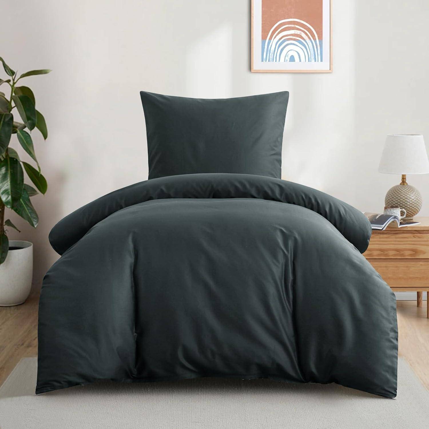 Ein ordentlich gemachtes Bett mit dunkelgrauer Bettwäsche, flankiert von einem Holznachttisch mit einer Lampe. Eine Pflanze und abstraktes Kunstwerk bereichern die Raumgestaltung.