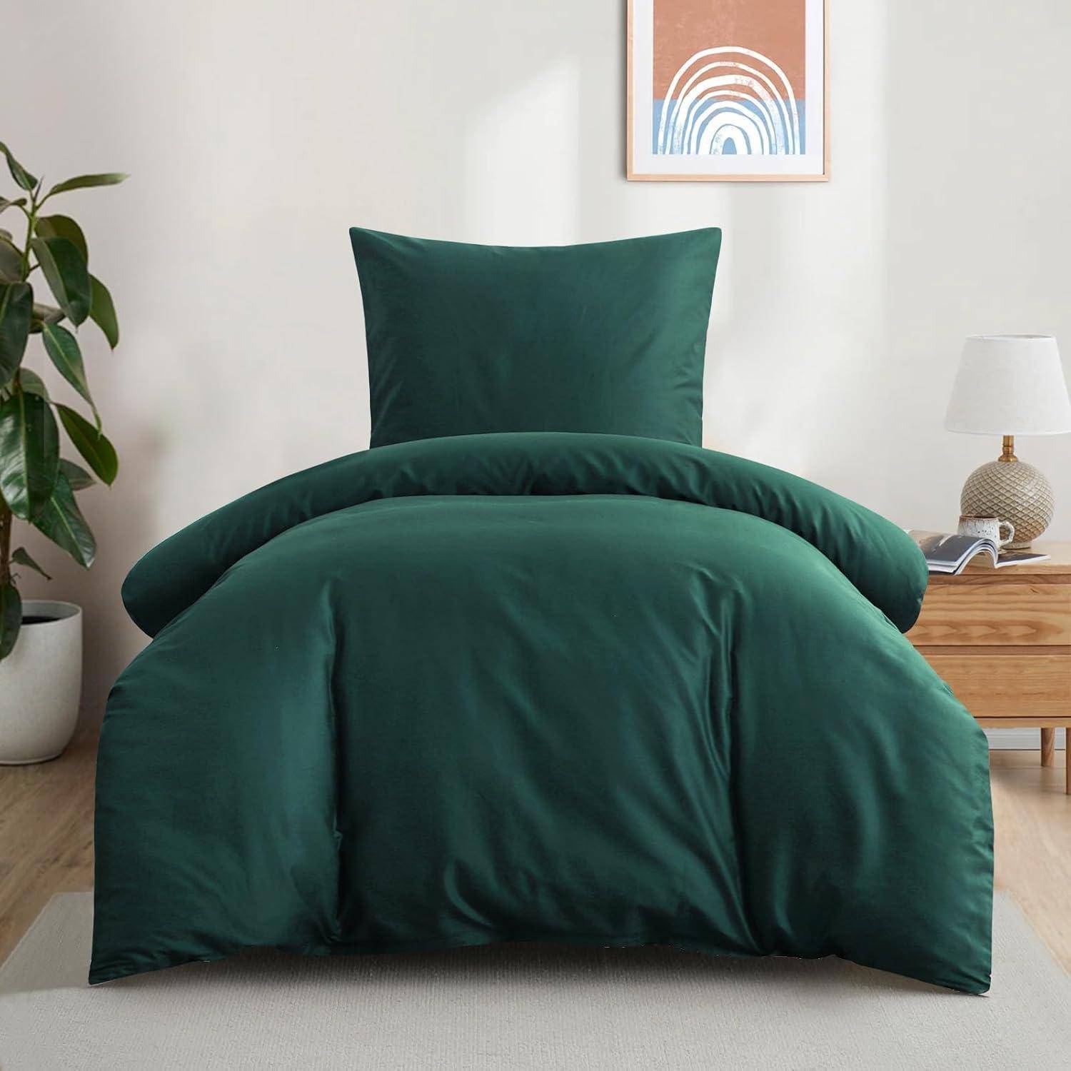 Ein ordentlich gemachtes Bett mit dunkelgrüner Bettwäsche steht in einem neutral gehaltenen Raum. Daneben befinden sich ein Nachttisch mit einer Lampe und einer dekorativen Pflanze.