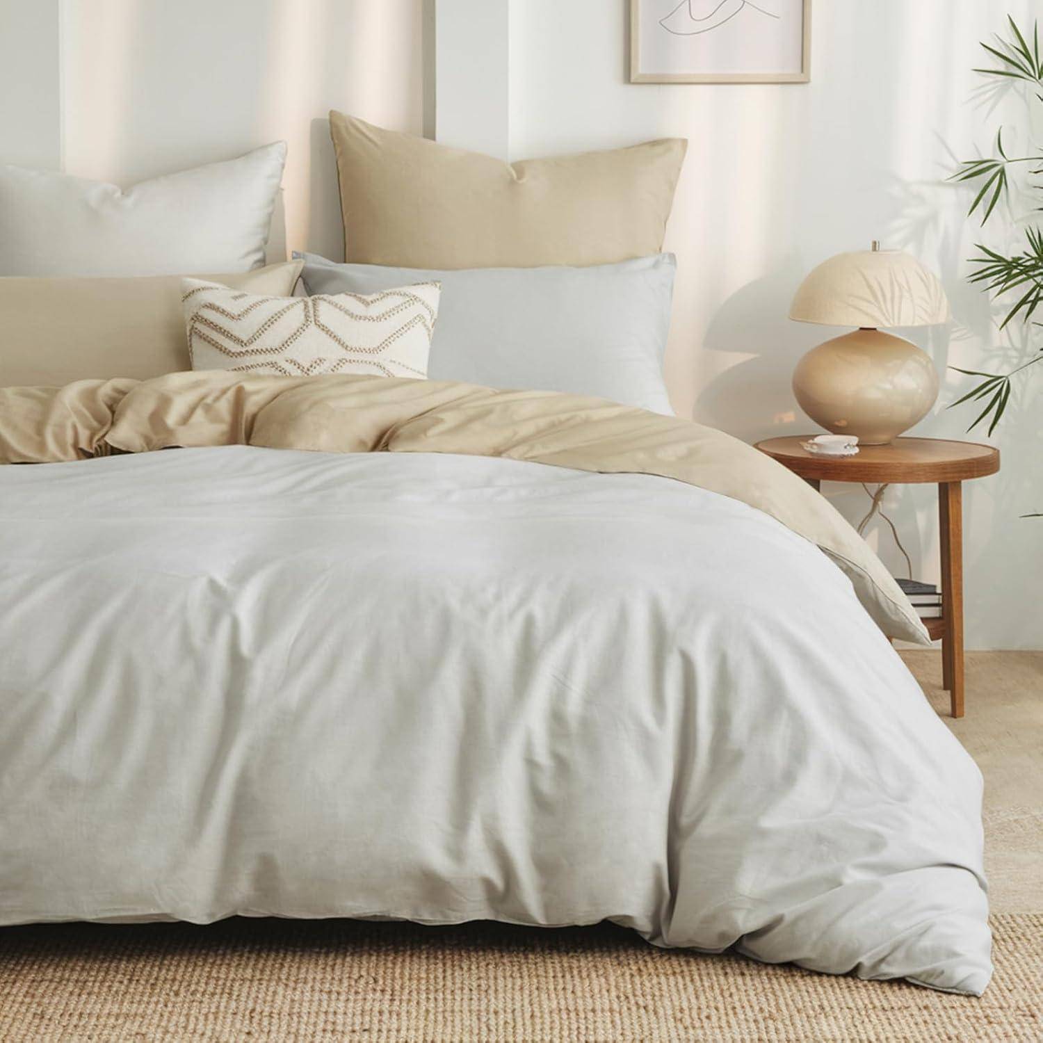 Ein gemütliches Schlafzimmer zeigt ein sorgfältig aufgemachtes Bett mit weißen und beigen Kissen sowie einer beigen Bettdecke. Ein Holznachttisch trägt eine Lampe und eine Vase.