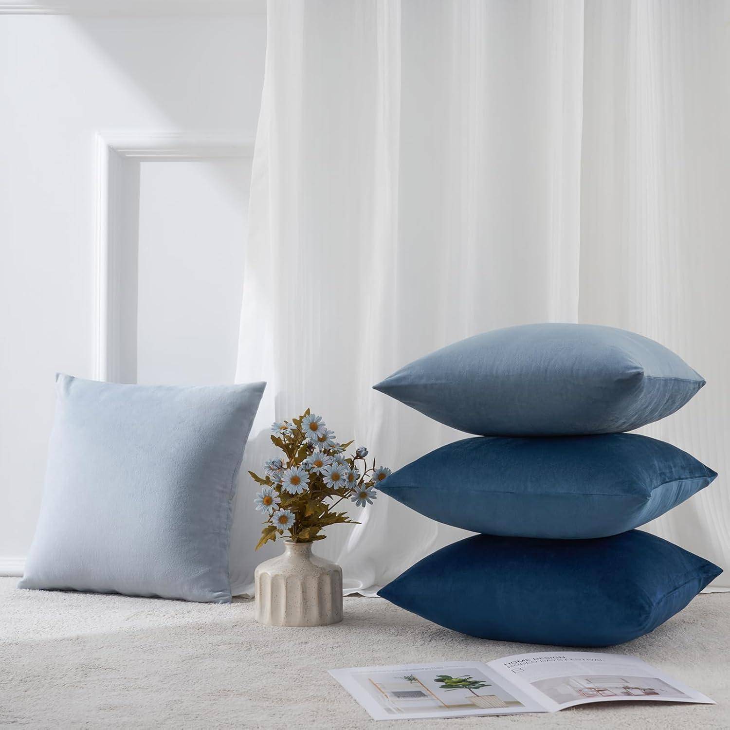 Ein Stapel von drei blauen Kissen neben einem hellblauen Kopfkissen auf einer weichen Oberfläche. In der Nähe eine kleine Vase mit Blumen und eine aufgeschlagene Zeitschrift.