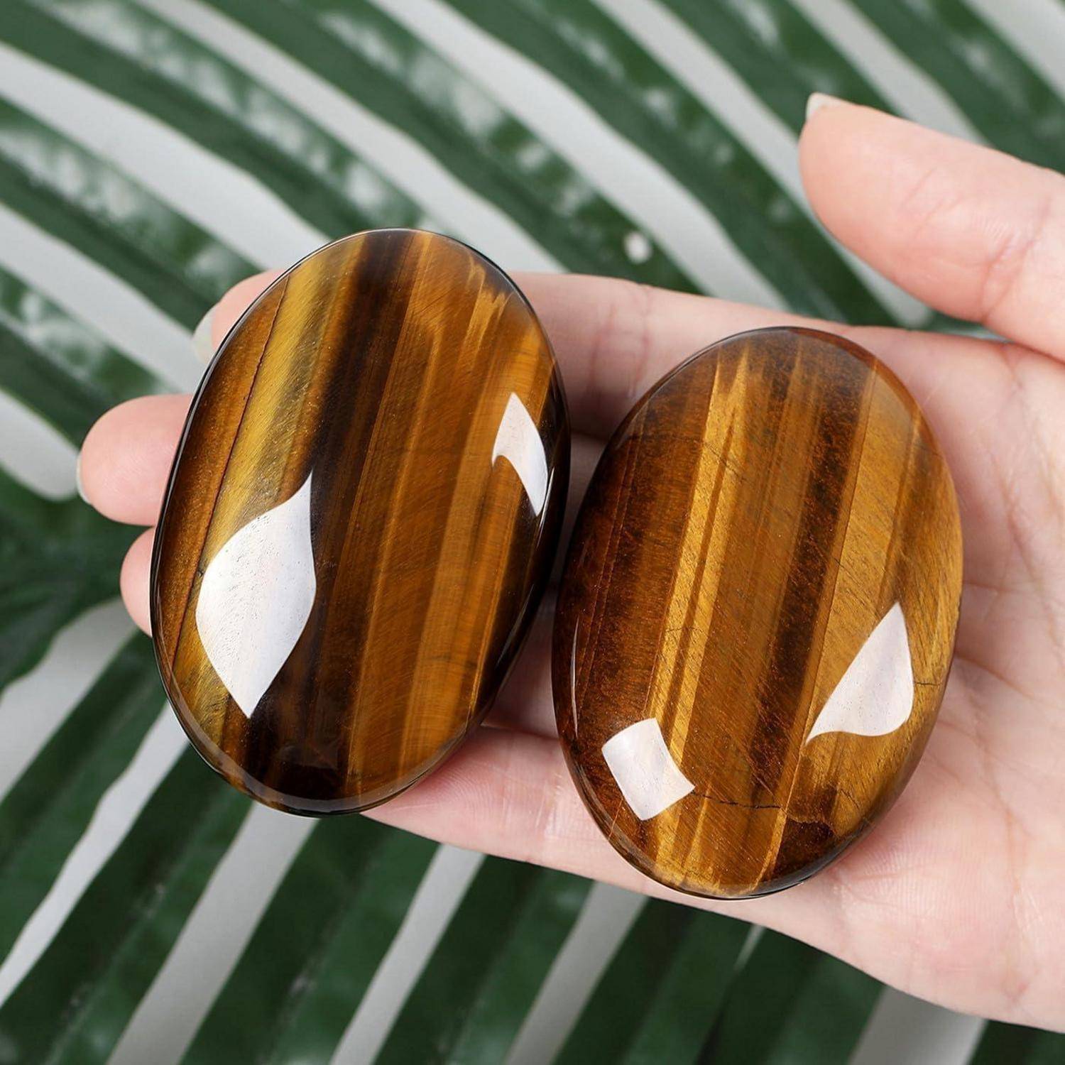Zwei ovale Tigerauge-Steine mit reflektierenden Oberflächen werden in einer Hand vor einem grün-weiß gestreiften Hintergrund gehalten.