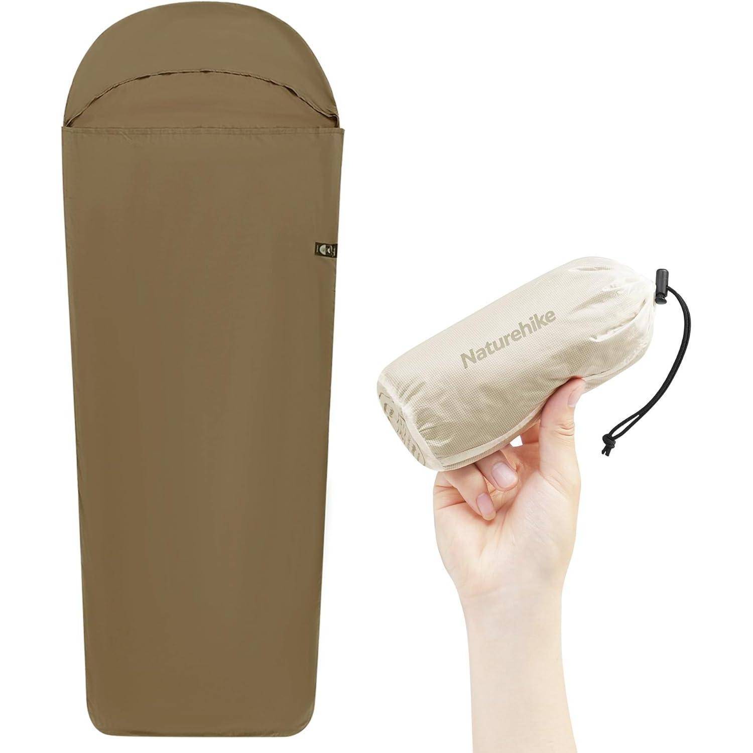 Eine kompakte, braune Schlafsack wird neben einer Hand gezeigt, die seinen kleinen, hellbeigen Packbeutel hält, was Tragbarkeit und Bequemlichkeit veranschaulicht.