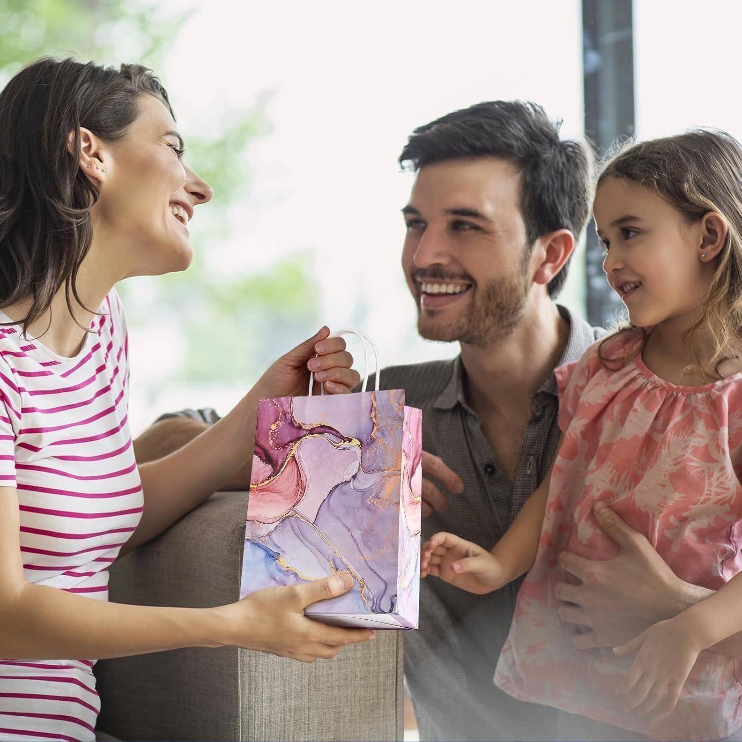 Eine Frau überreicht einem lächelnden Mann, der ein junges Mädchen trägt, eine Geschenktasche, was auf eine Feier oder einen besonderen Anlass in einer Wohnumgebung hindeutet.