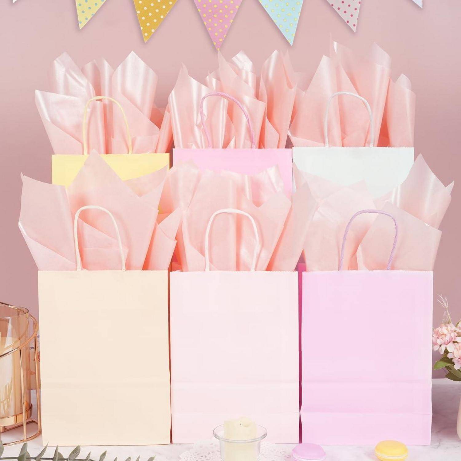Rosa und gelbe Geschenktüten mit Seidenpapier auf einem Tisch, unter pastellfarbener Wimpelkette.