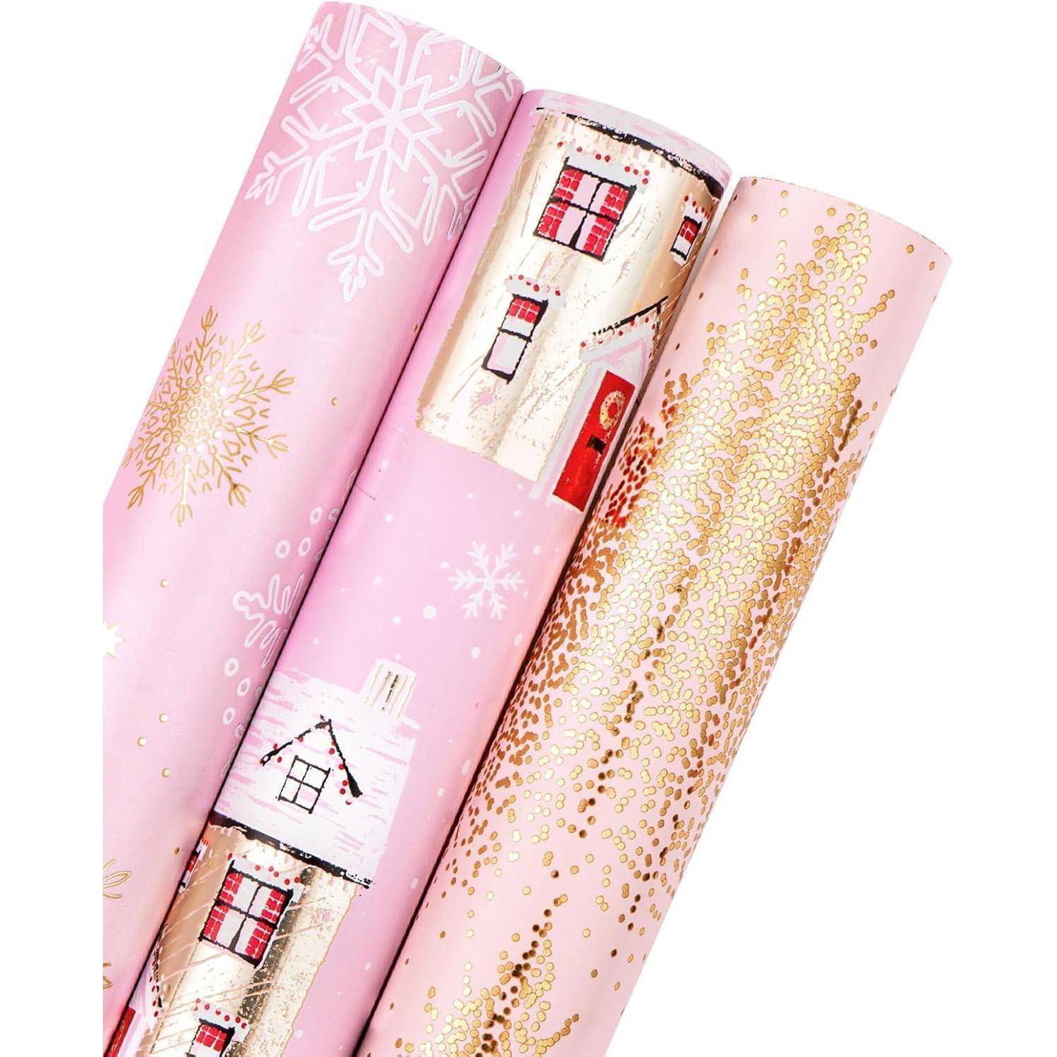 Drei Rollen rosa Weihnachtsgeschenkpapier mit Schneeflocken, Häusern mit roten Türen und Fenstern sowie goldenen Verzierungen.