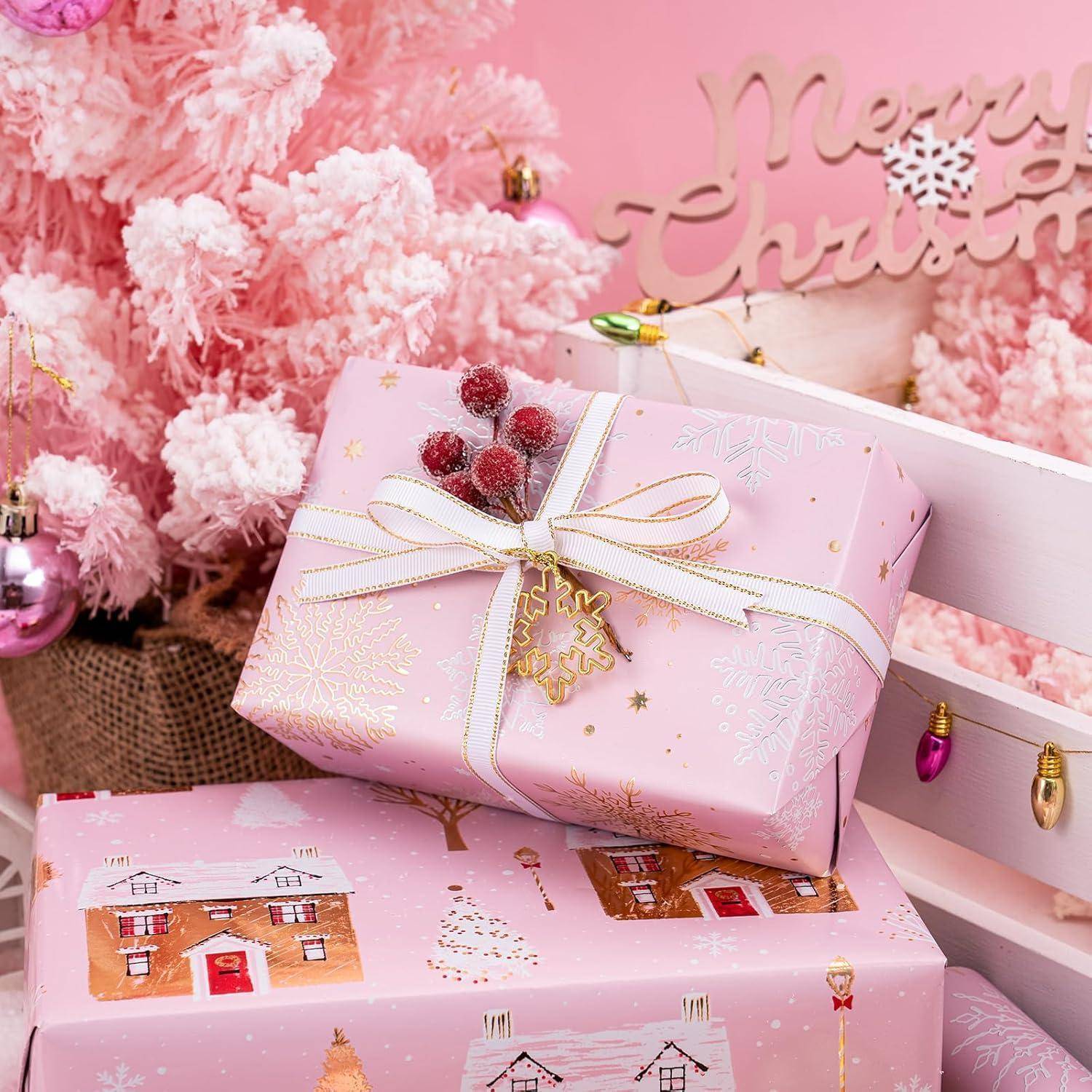Eine rosa Geschenkbox mit einem festlichen Band liegt neben einem rosa Weihnachtsbaum, der mit Christbaumschmuck verziert ist. Ein Schild mit der Aufschrift "Frohe Weihnachten"" befindet sich im Hintergrund.