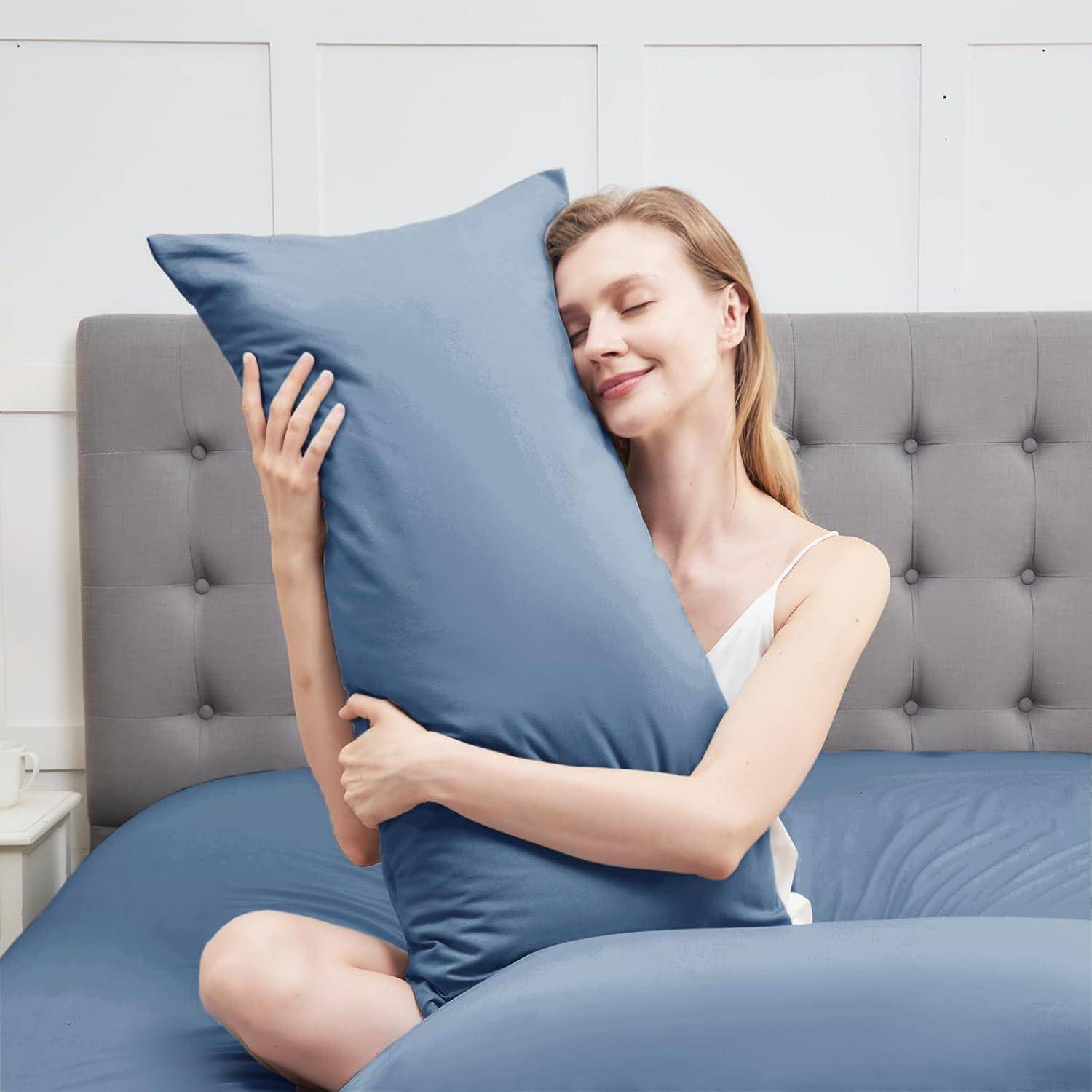 Eine Person umarmt glücklich ein großes blaues Kissen und sitzt dabei auf einem Bett mit grauem Kopfteil, wodurch eine entspannte und zufriedene Stimmung entsteht.