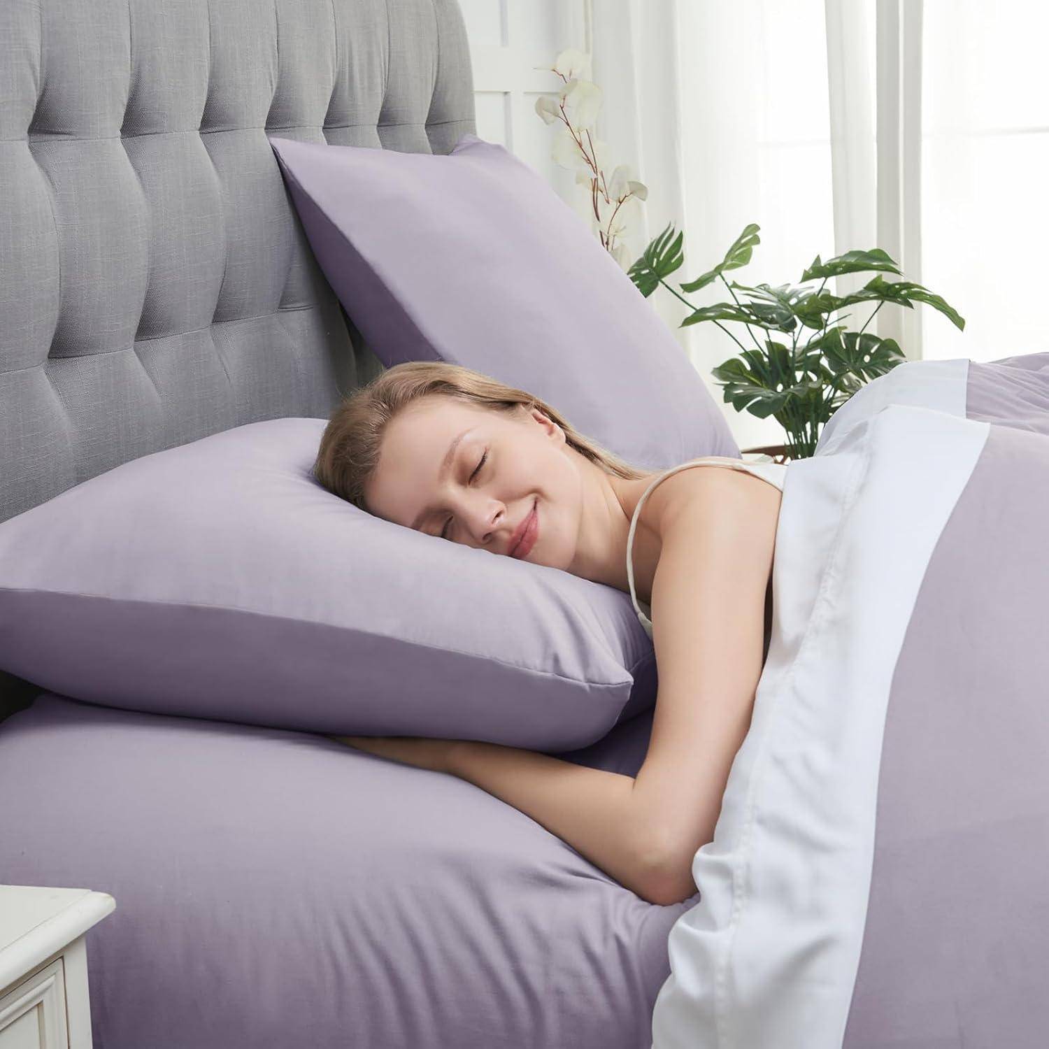 Eine Frau schläft friedlich in einem Bett mit Lavendel-Bettwäsche und -Kissen und lächelt, während im Hintergrund eine kleine Pflanze steht.