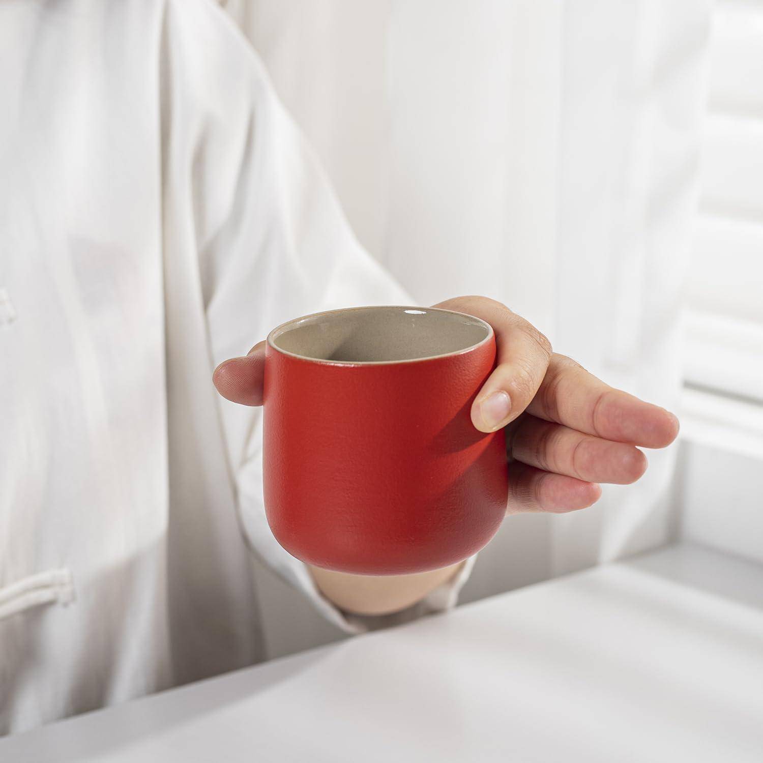 Eine Hand hält eine kleine, rote Keramiktasse. Der Hintergrund zeigt eine verschwommene, helle Umgebung.