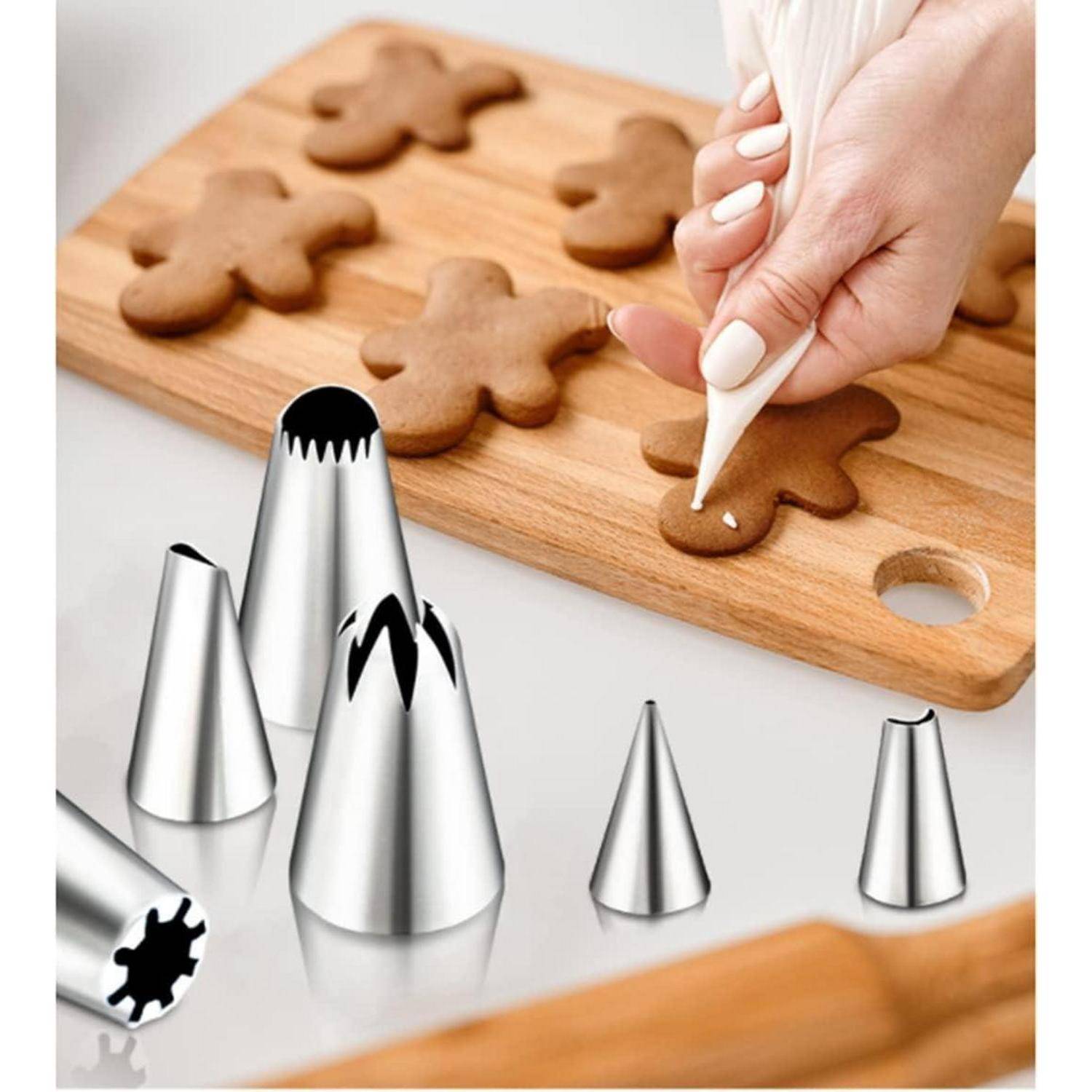 Eine Hand verziert Lebkuchenplätzchen auf einem Holzbrett mit einem Spritzbeutel. Verschiedene Metallspritztüllen sind auf einer weißen Oberfläche verstreut.
