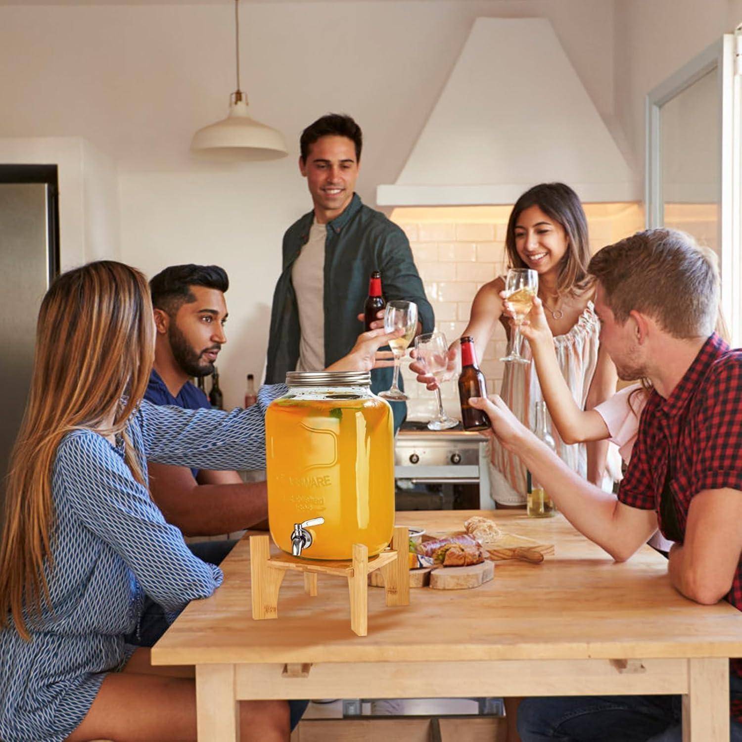 Sechs Freunde in einer Küche, die lächeln und mit Getränken anstoßen. Ein Getränkespender und Snacks stehen auf dem Tisch und sorgen für eine festliche Atmosphäre.